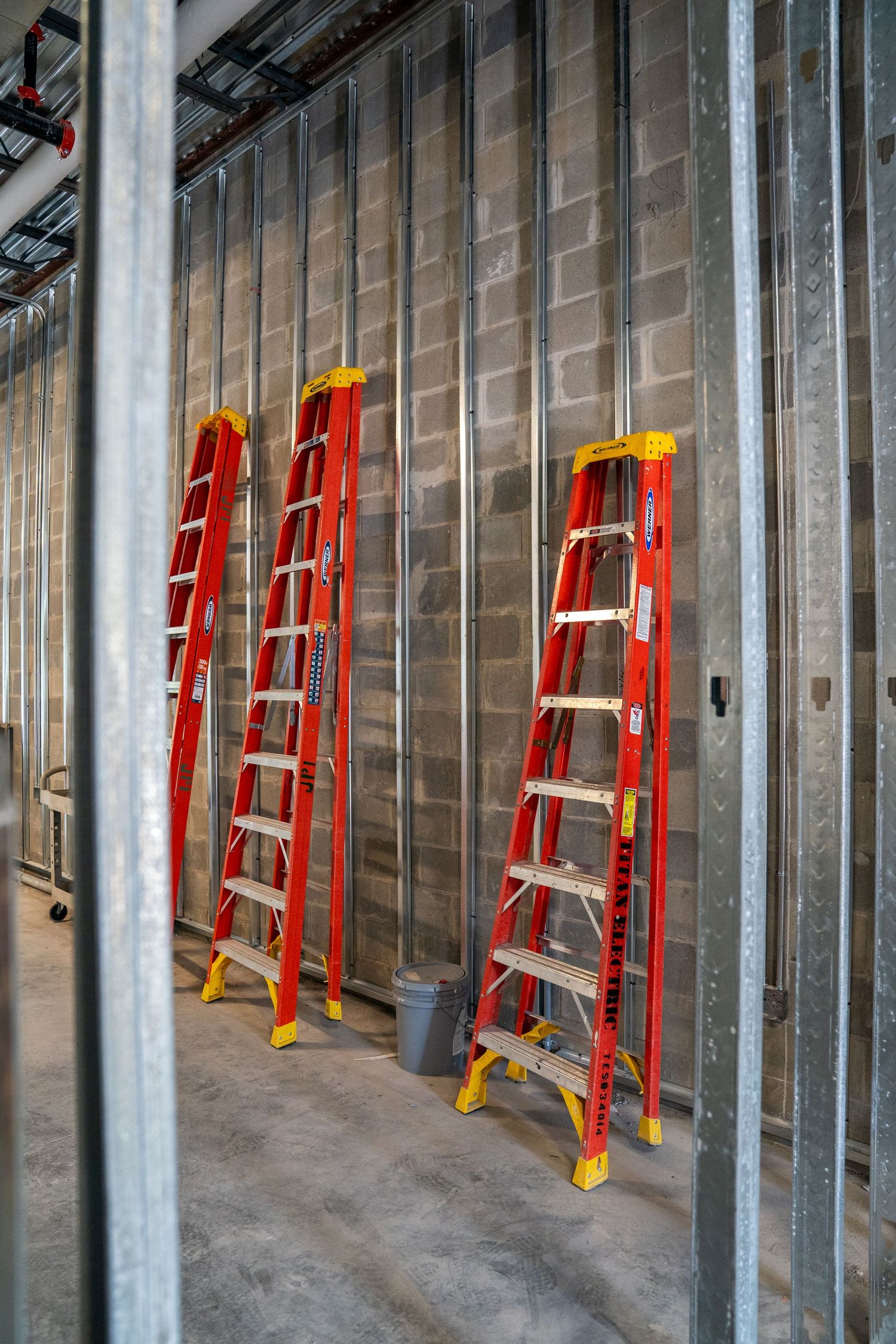 Row of red ladders leaning against metal wall studs in commercial construction site.