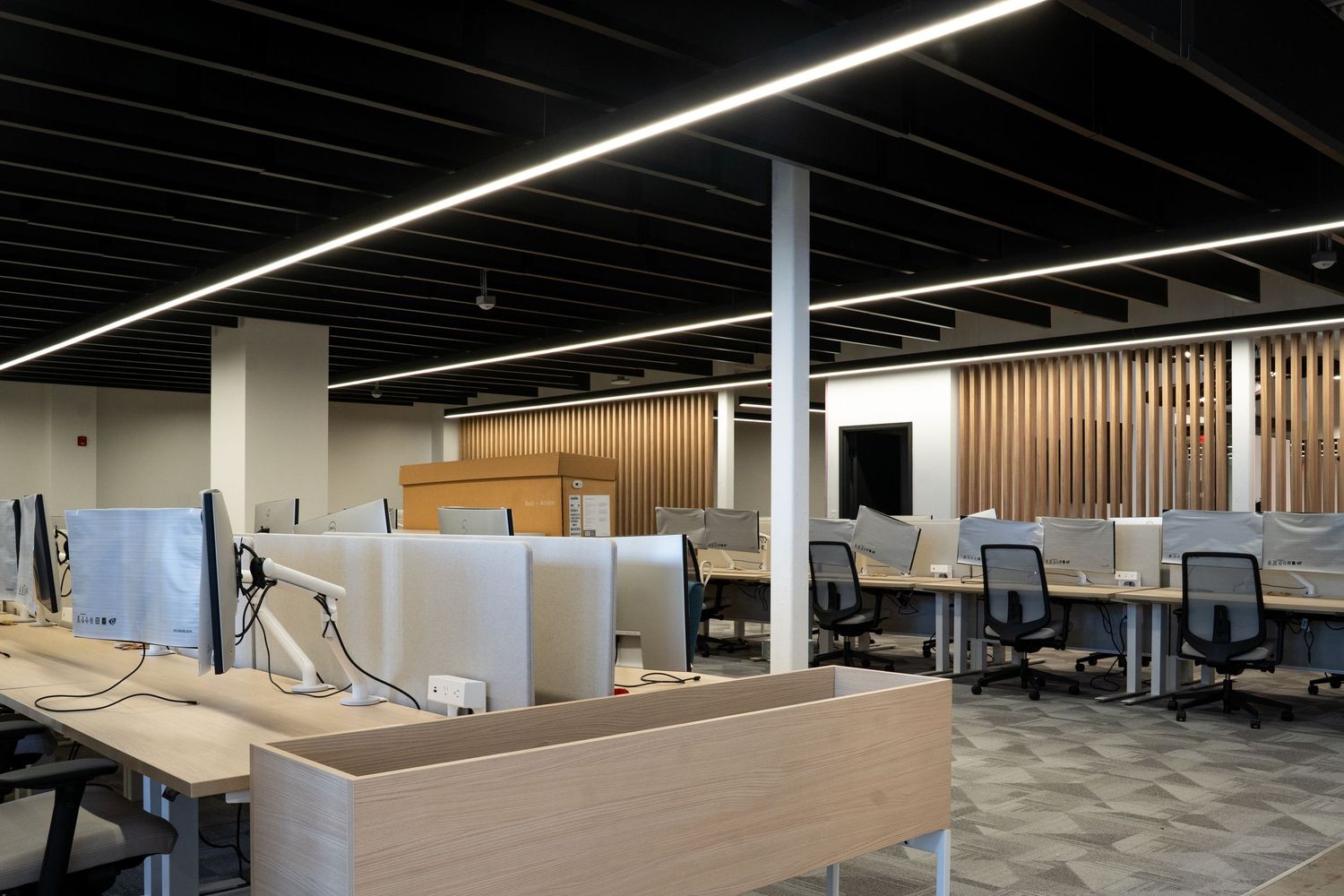 Modern open office space with light wood desks, gray dividers, and white columns under linear lighting fixtures.