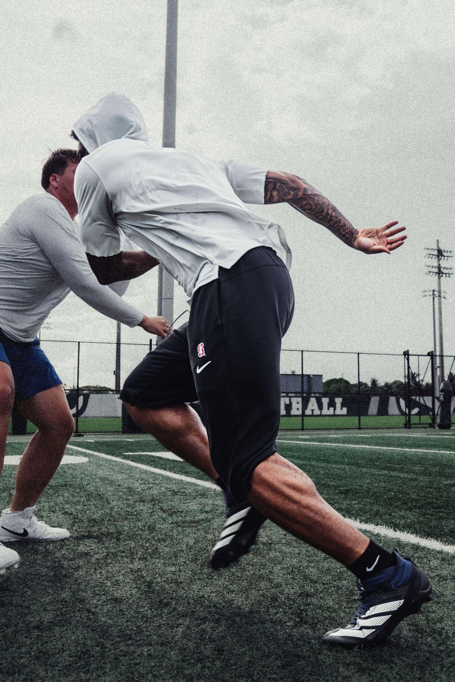 Two football players practice pass rush drills on a turf field, one with tattoos wearing a hoodie and Adidas cleats.