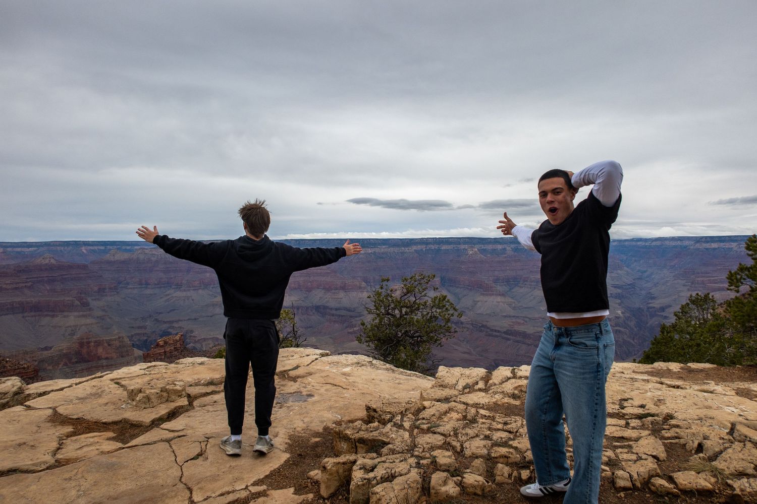 Two people strike poses on a rocky overlook at the Grand Canyon with dramatic cloudy skies.