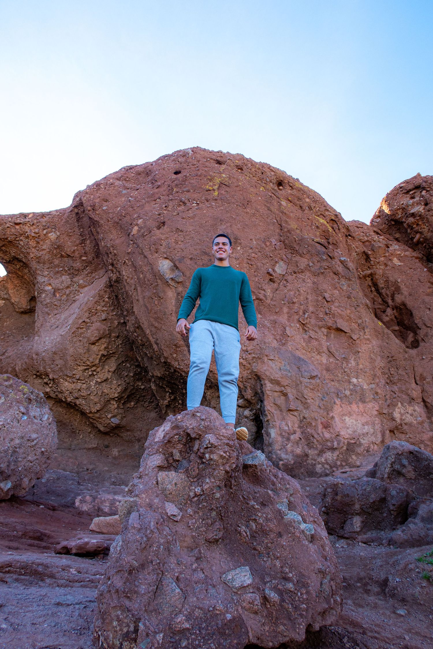 A person in a blue sweater stands on red rock formations against a bright blue sky.
