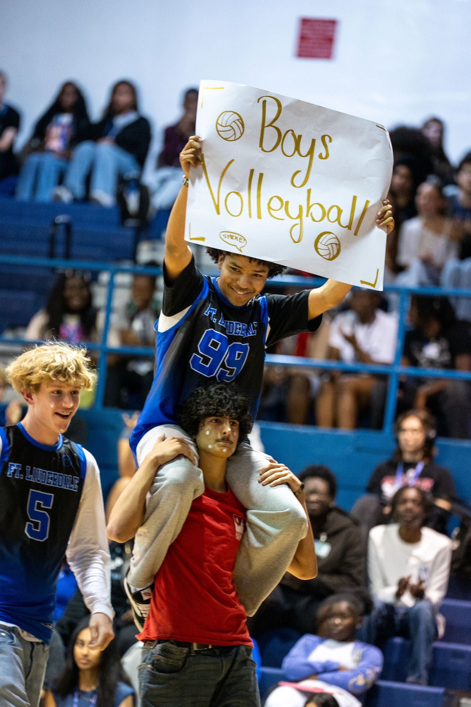 Student holds Boys Volleyball sign while sitting on teammate's shoulders at a high school pep rally in a gymnasium.