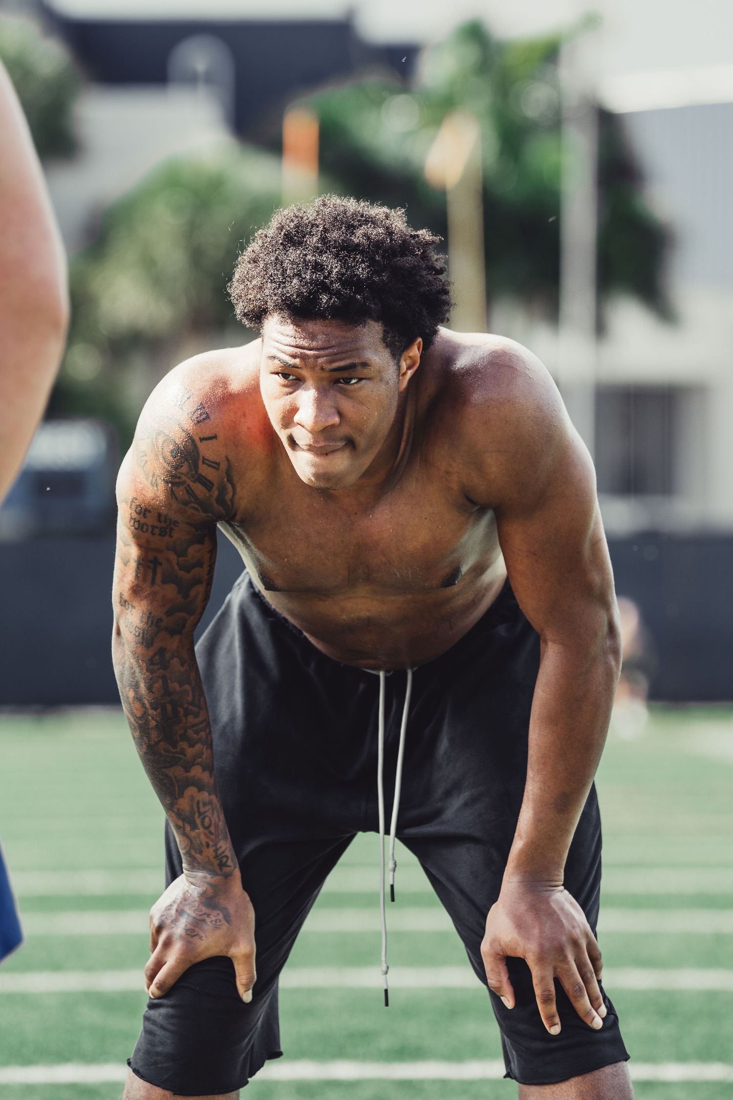 Muscular shirtless athlete with tattoos resting hands on knees on a green turf football field.
