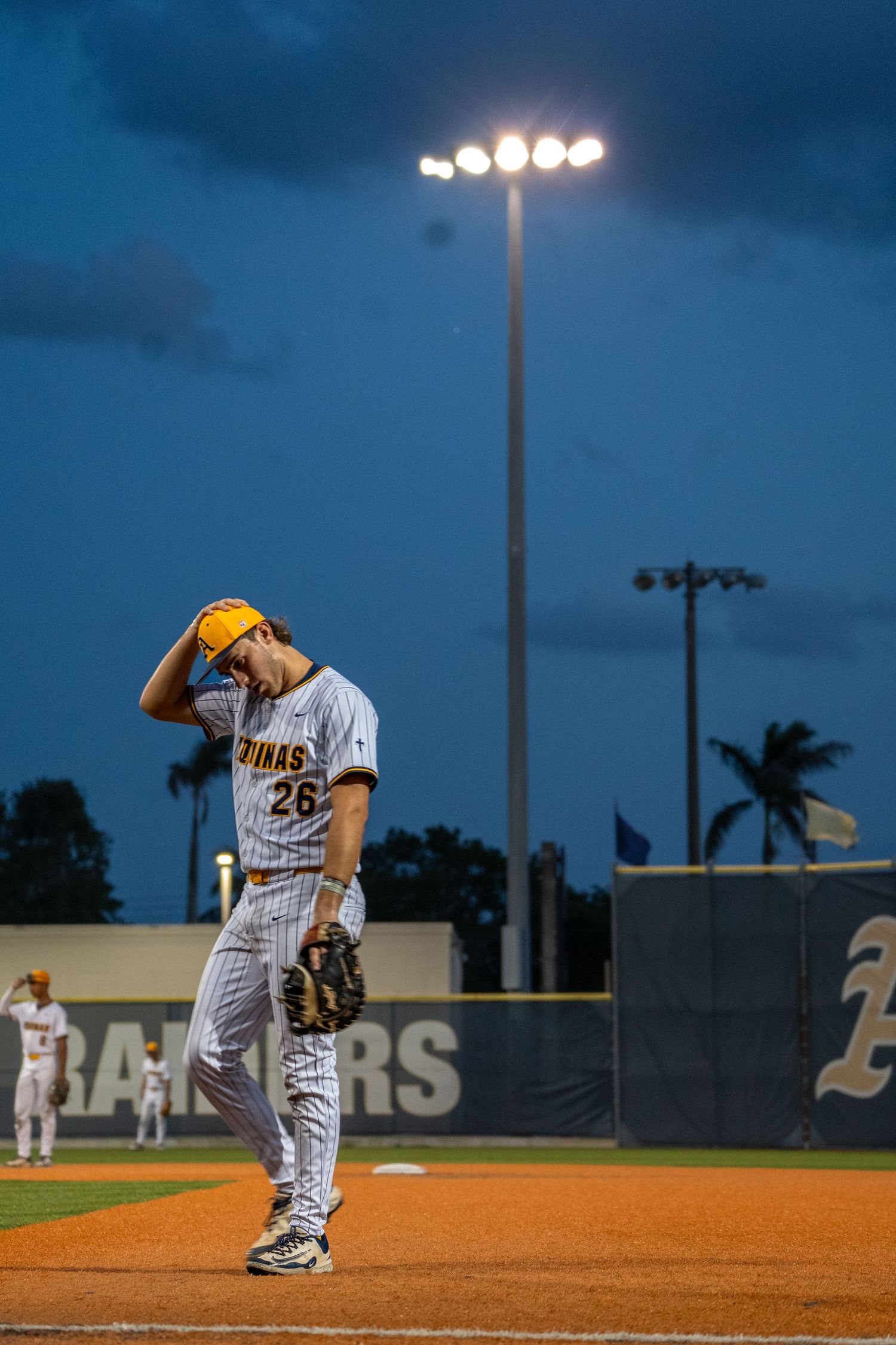 Baseball players on a field during an evening game with stadium lights illuminating the dark blue sky.