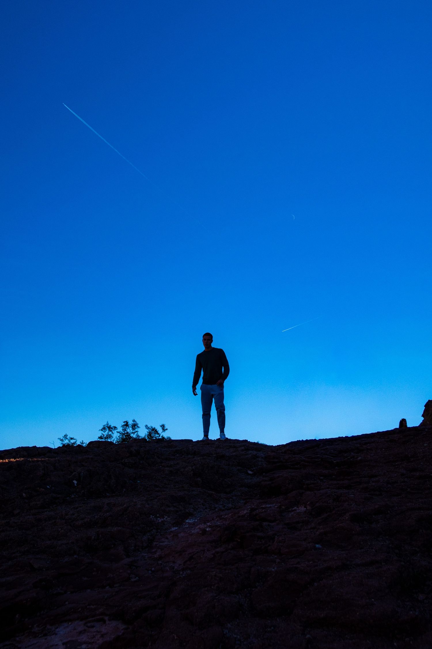 Silhouette of a person standing on a hill against a vibrant blue sky with contrails.