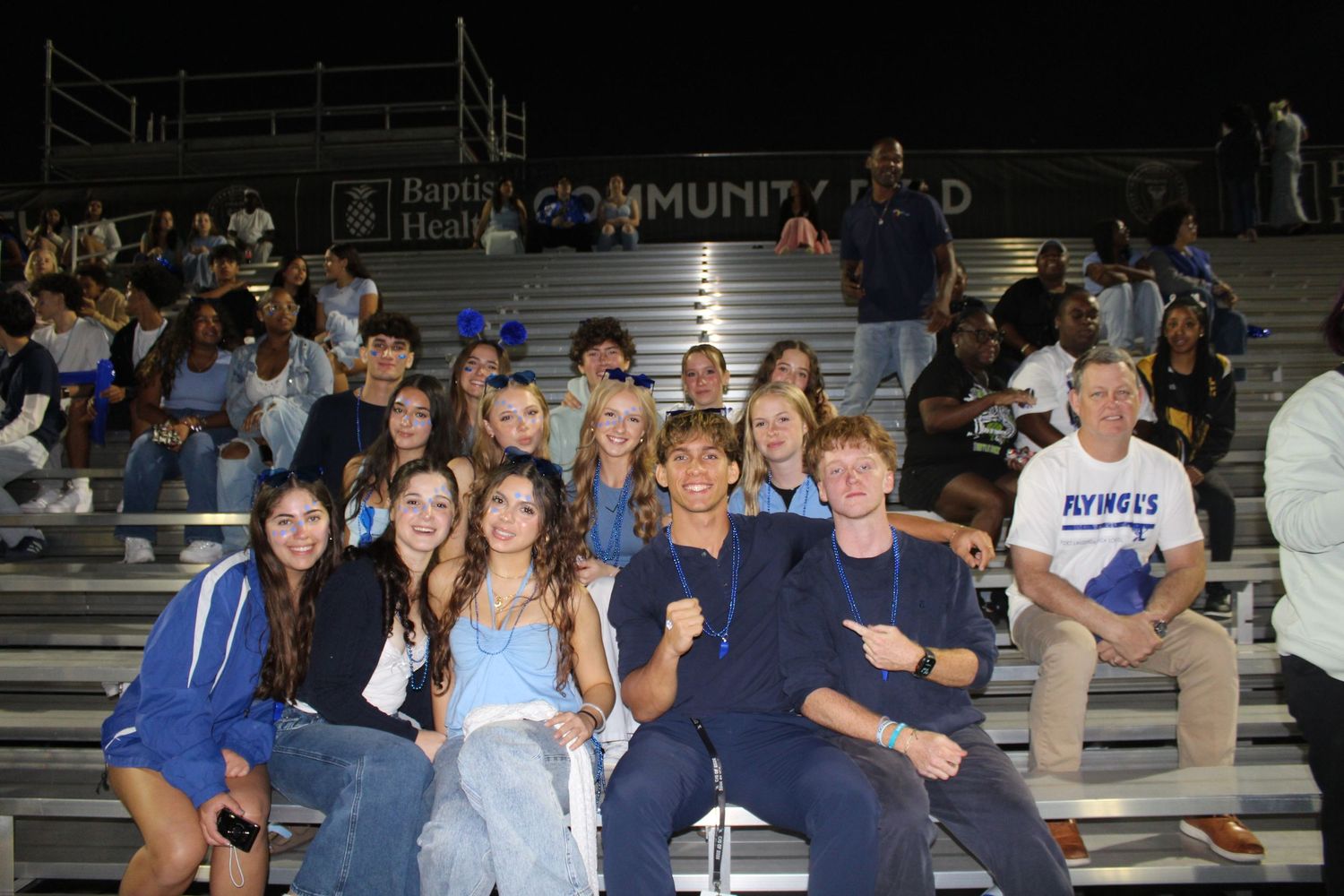 A group of students sitting together in bleachers at a nighttime sporting event.