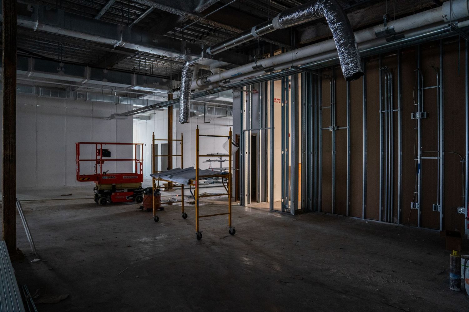 Interior construction site with metal framing, scissor lift, and exposed mechanical systems overhead.