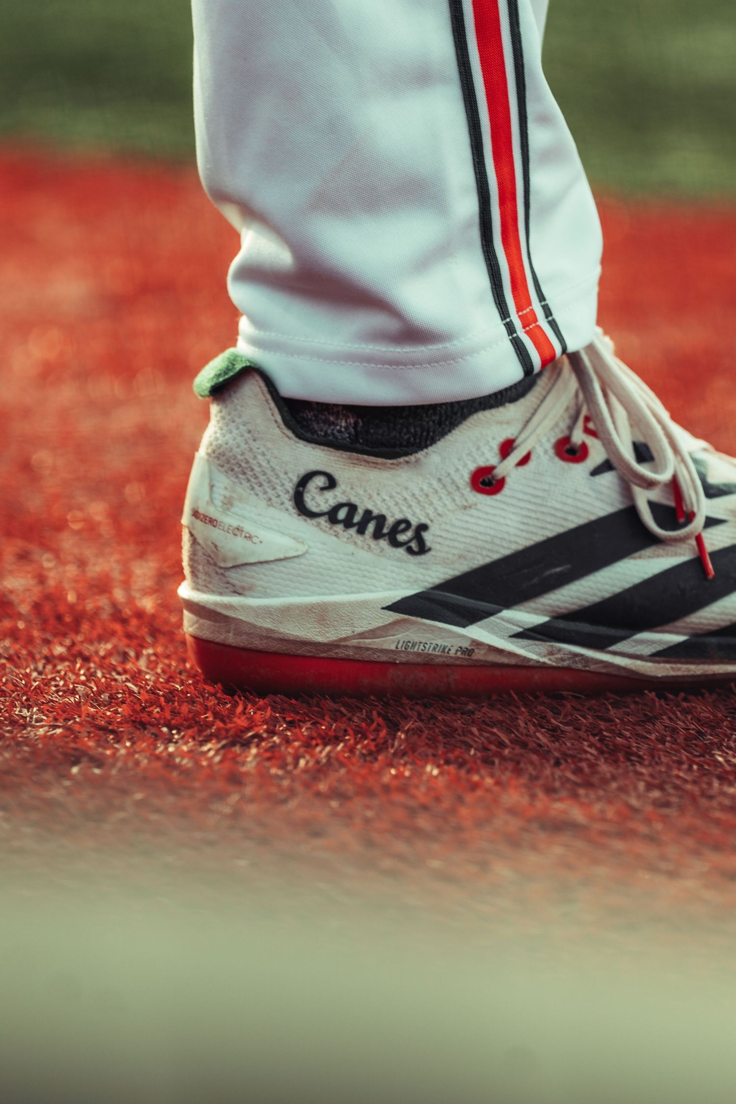 Close-up of white Canes baseball cleats with red and black stripes on red clay field.