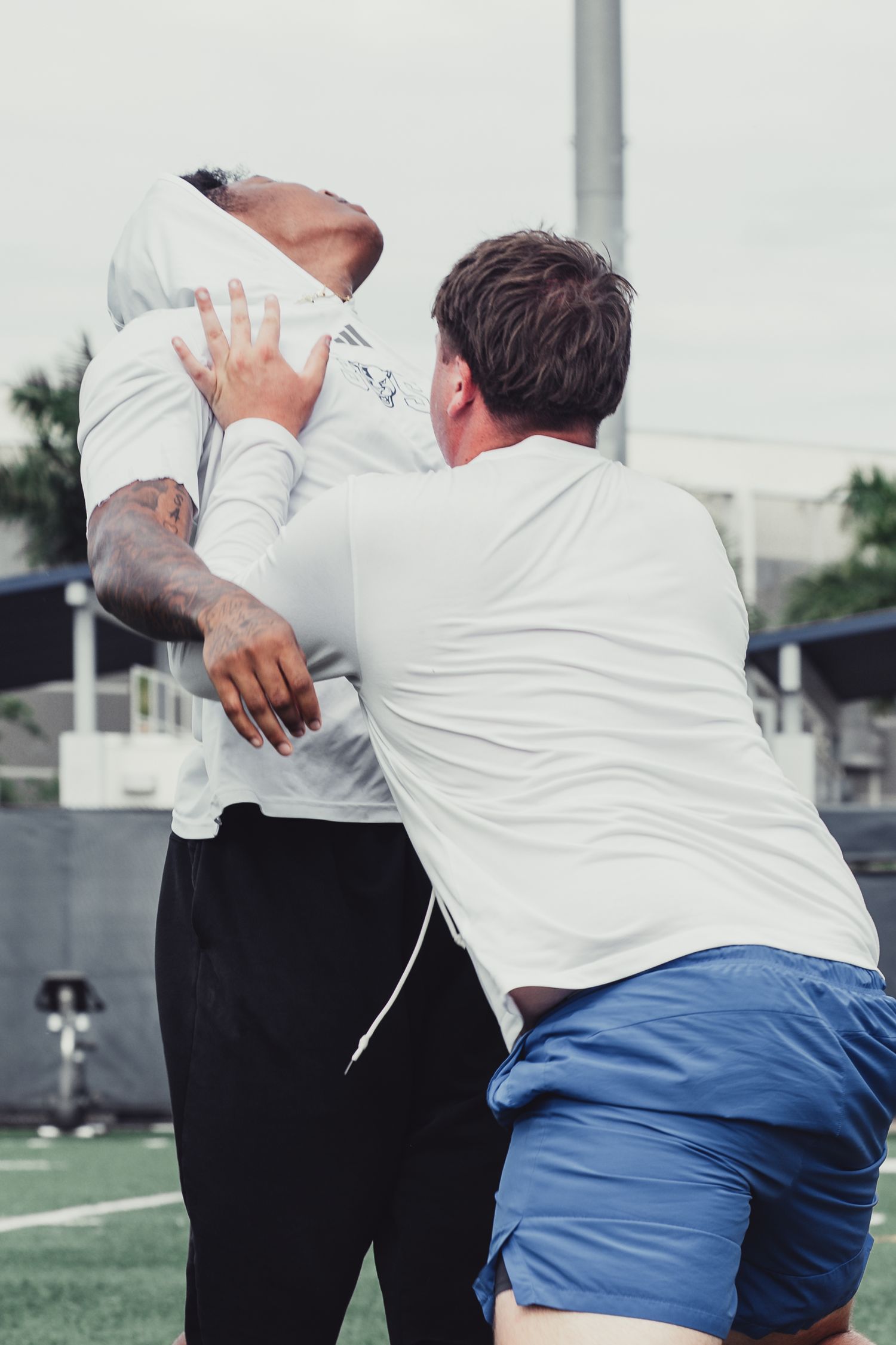Two athletes in white shirts practice football blocking drills on a green field outdoors.