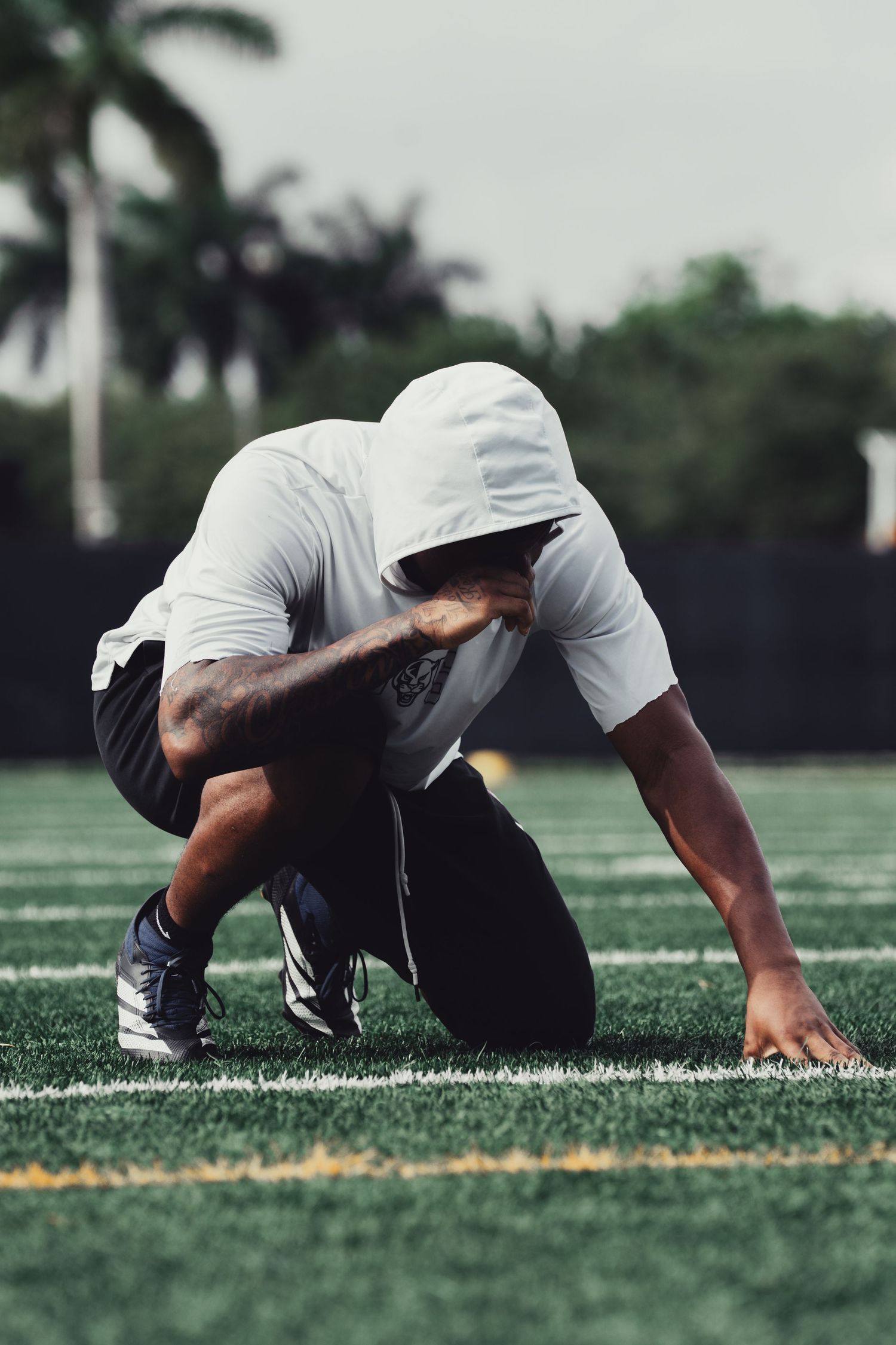 Athlete in white hoodie crouches in starting position on green turf field with palm trees in background.