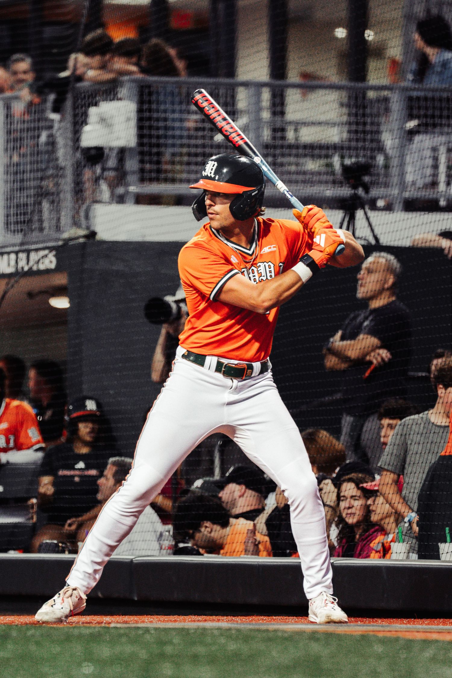 Baseball player in orange uniform stands ready at bat during a night game with crowd in background.