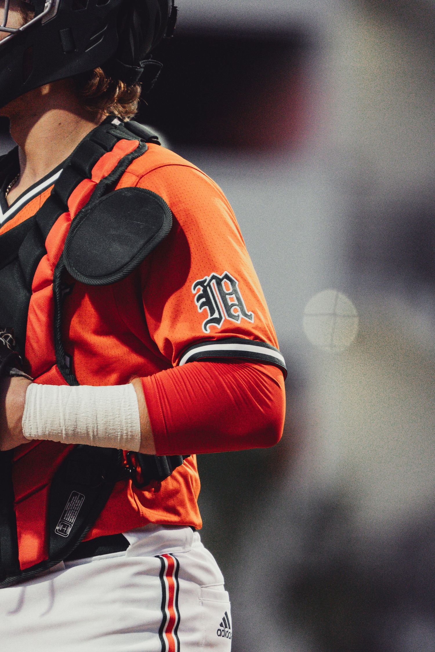 A baseball catcher in an orange Adidas uniform with an M logo stands ready during a game.
