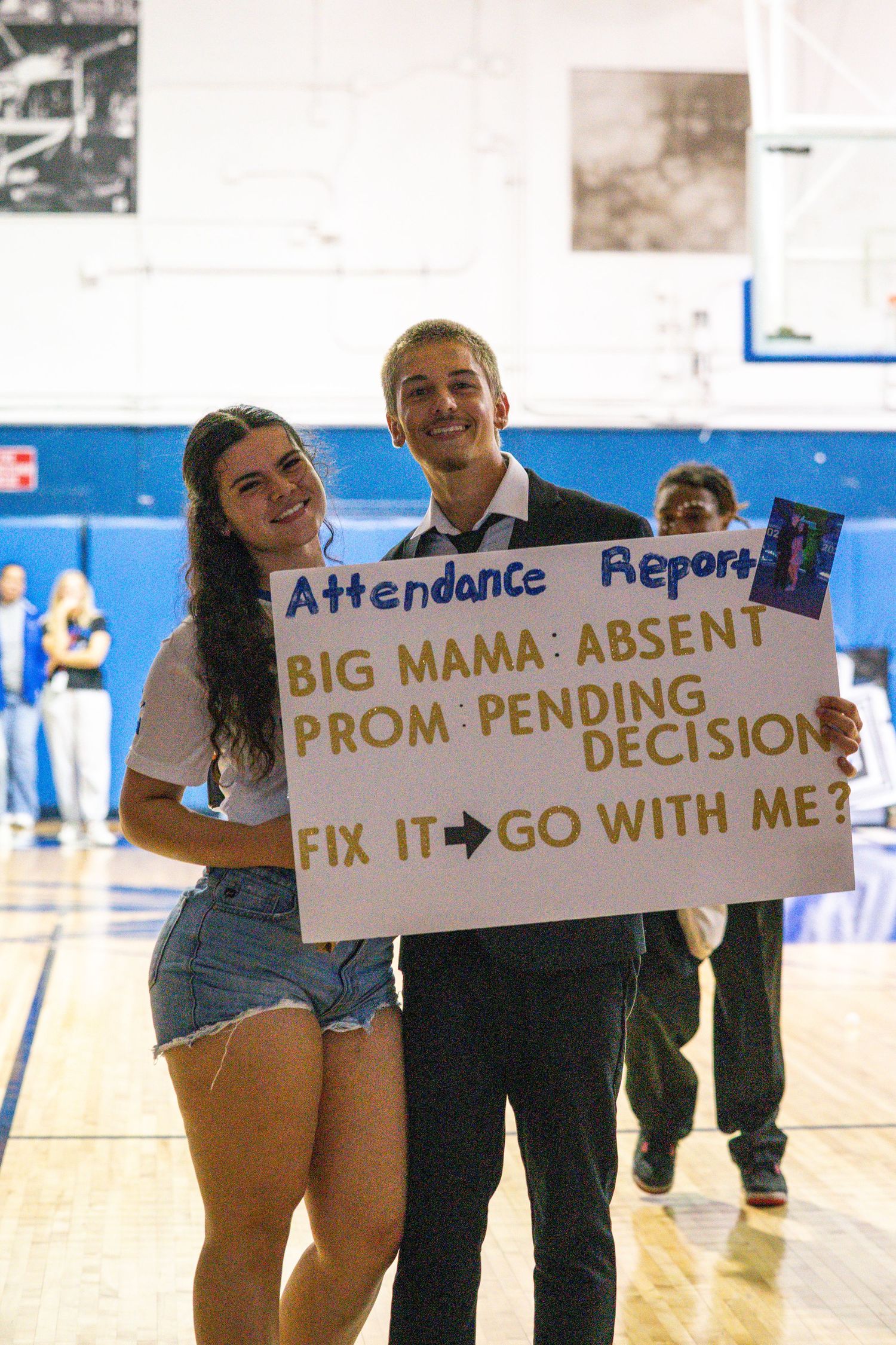 Teen boy holds promposal sign reading Attendance Report asking Big Mama to prom in school gymnasium.