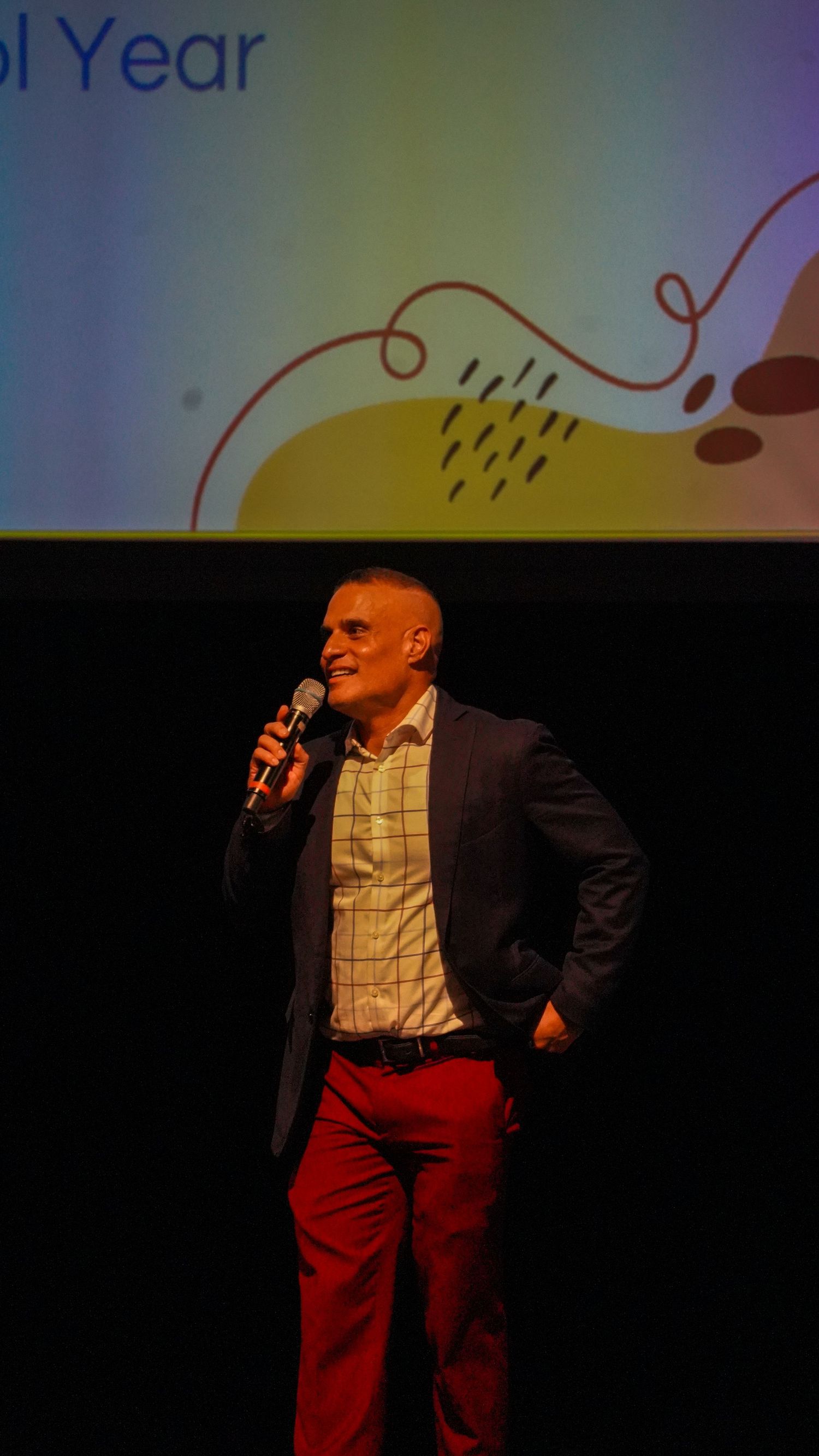 Speaker in business attire presents on stage with colorful projection behind.