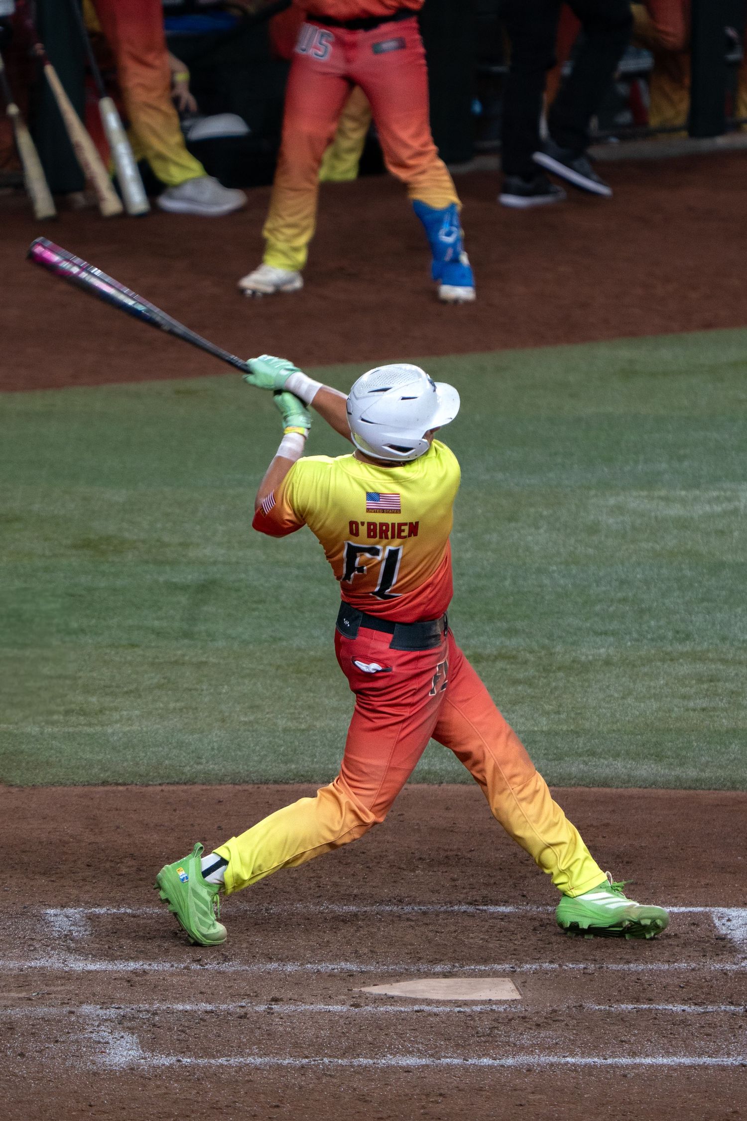 Baseball player in orange and yellow gradient uniform swinging bat at home plate.