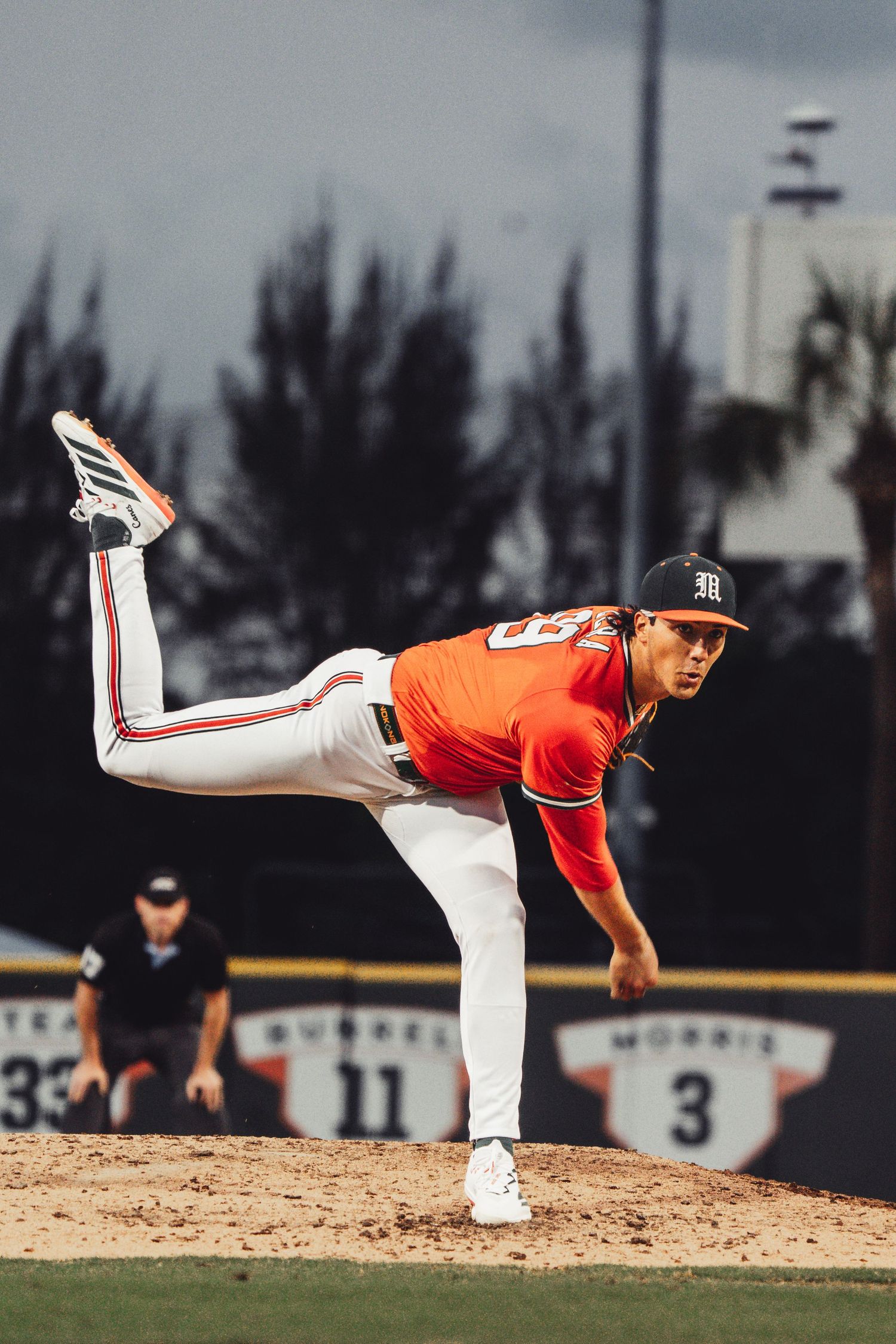A baseball pitcher in an orange uniform delivers a pitch on a sunny outdoor baseball field.