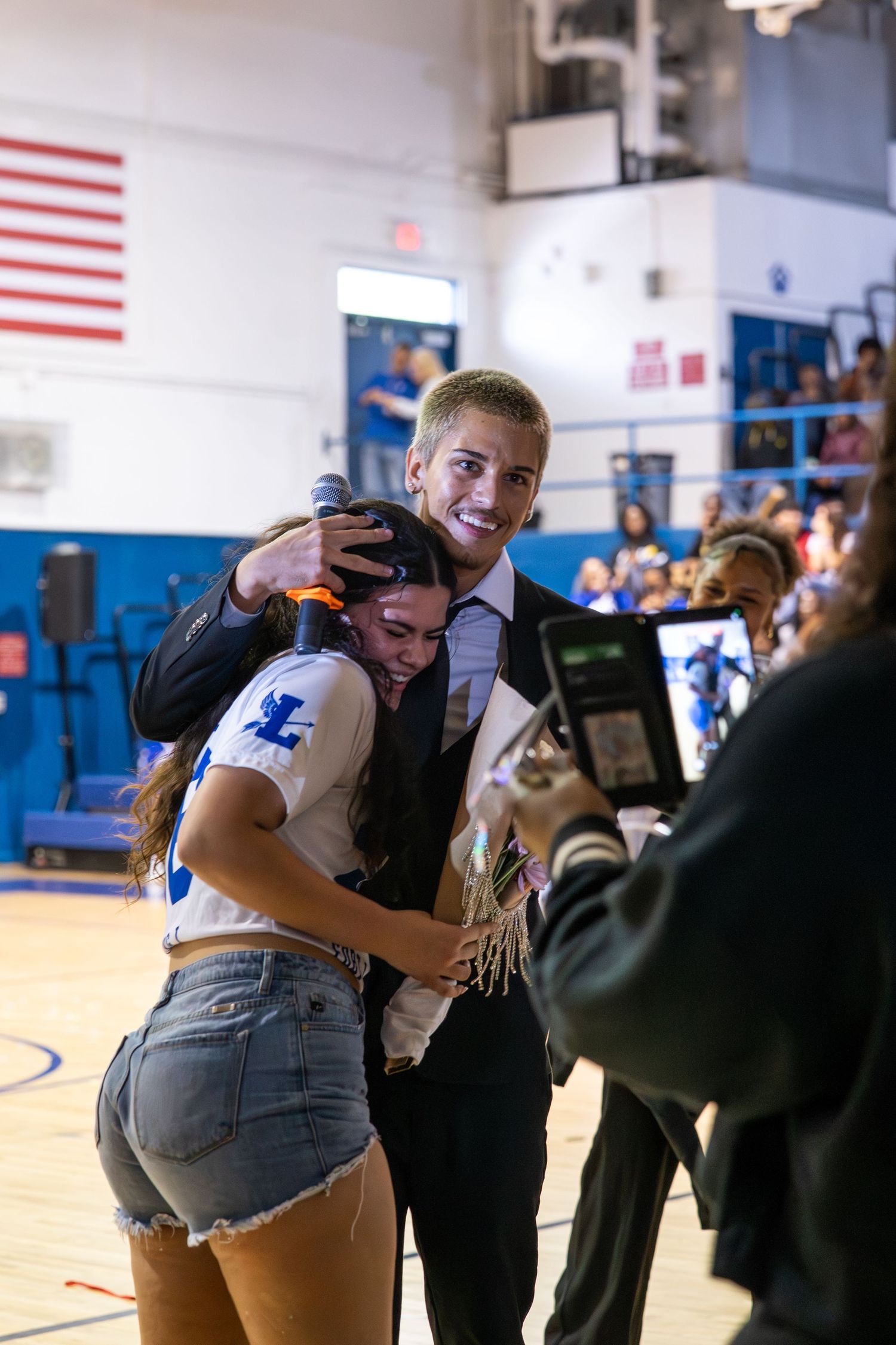 A smiling man hugs a young woman in a school gymnasium during a celebratory event, with onlookers nearby.
