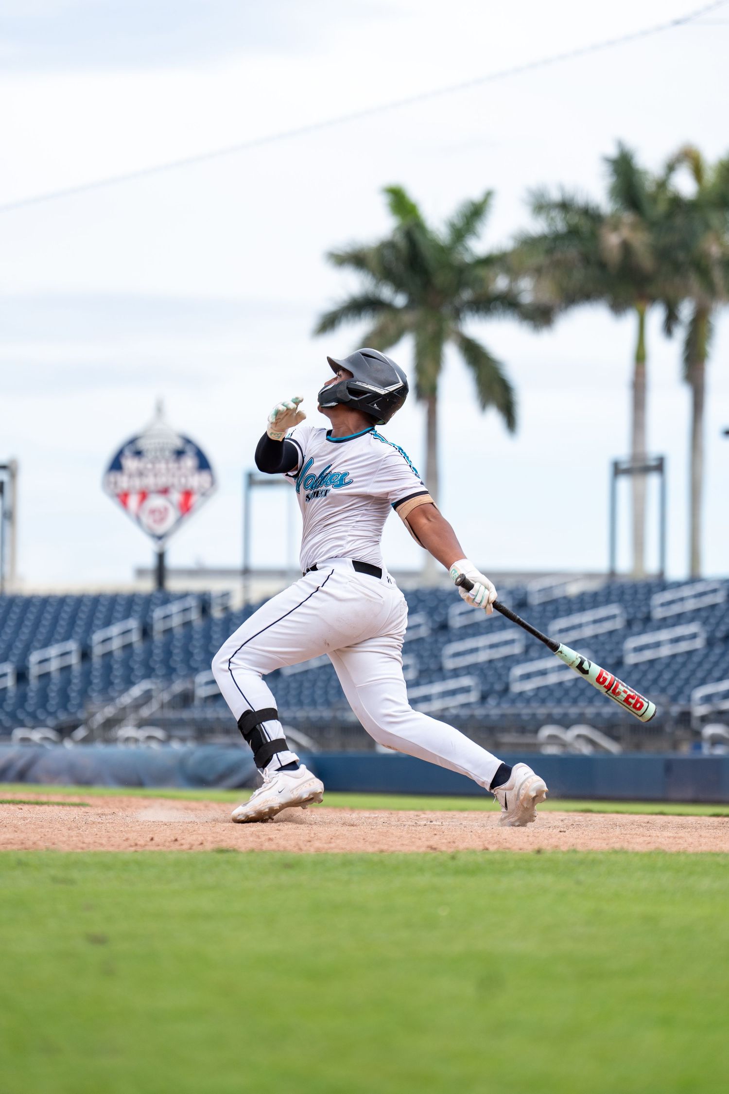 Baseball player in white uniform follows through on a powerful swing at a professional stadium with palm trees.