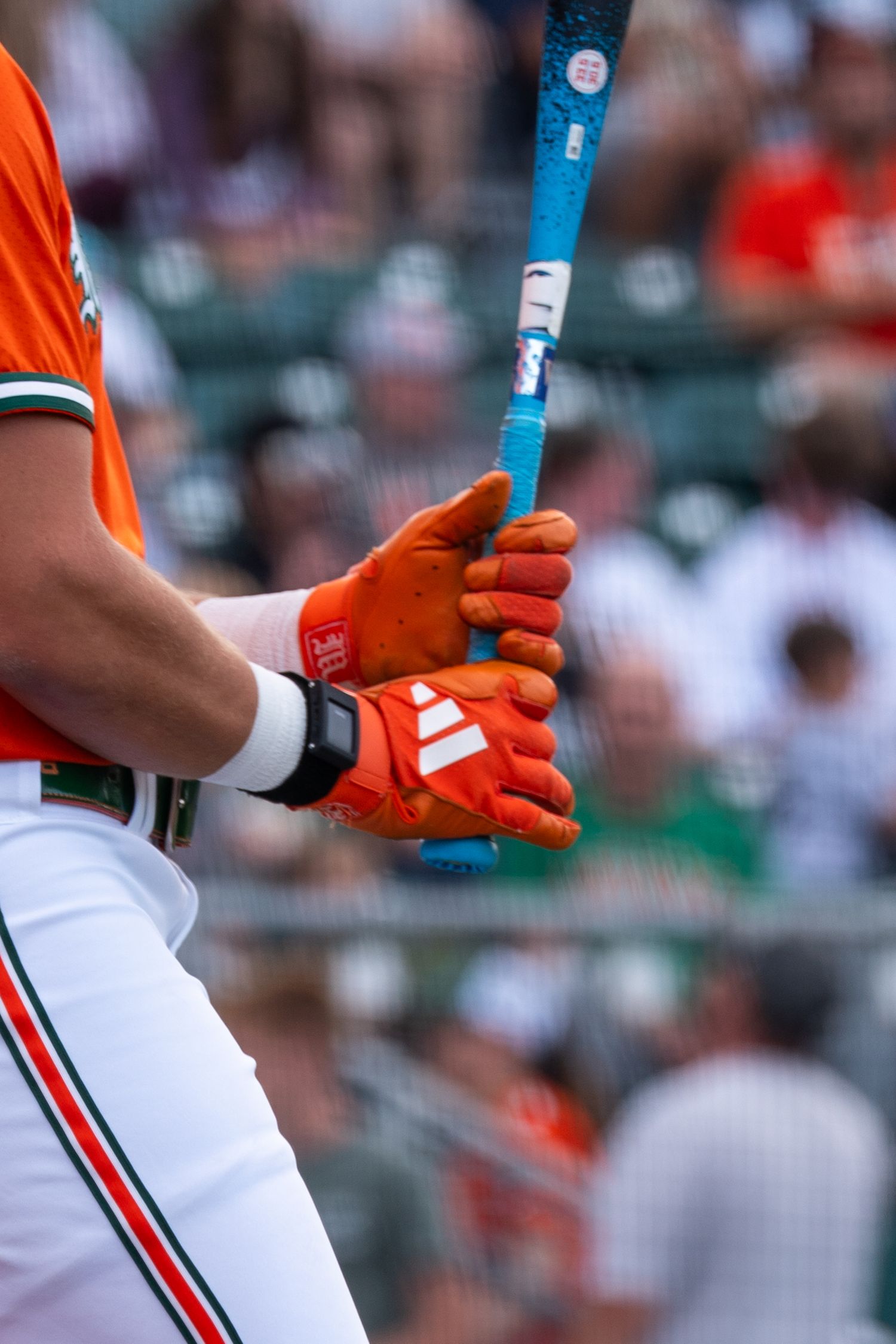 Close-up of a baseball batter gripping a blue bat with orange Adidas batting gloves during a game.