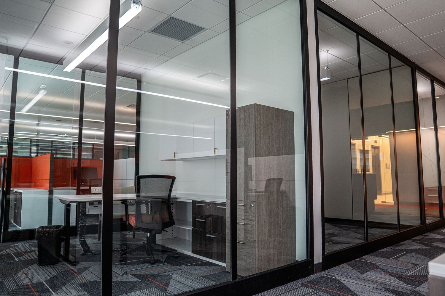 Modern office workspace with glass walls, ergonomic furniture, and geometric carpet patterns.