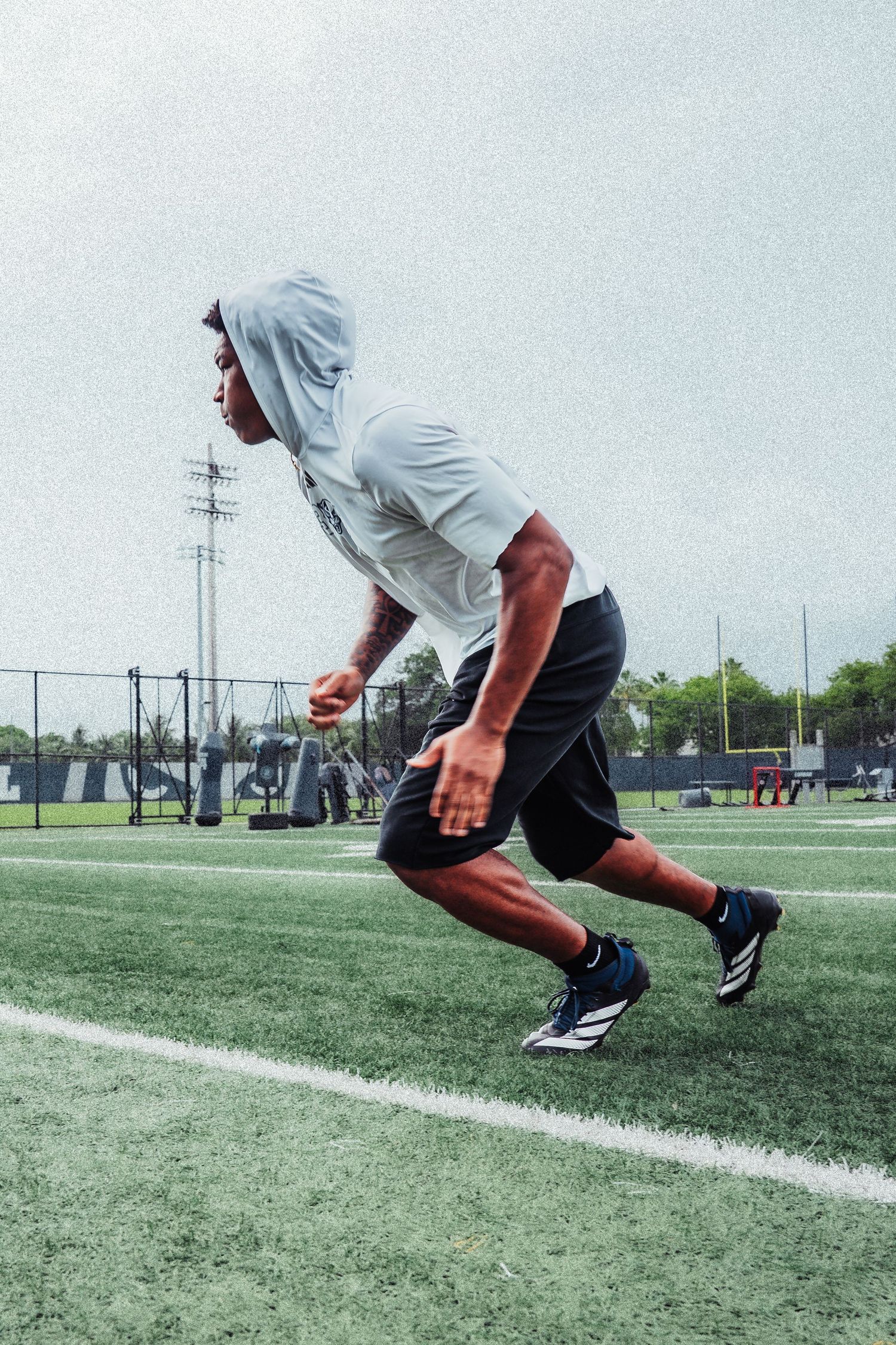Athlete in a gray hoodie and black shorts sprints across a green turf football field wearing blue Adidas cleats.