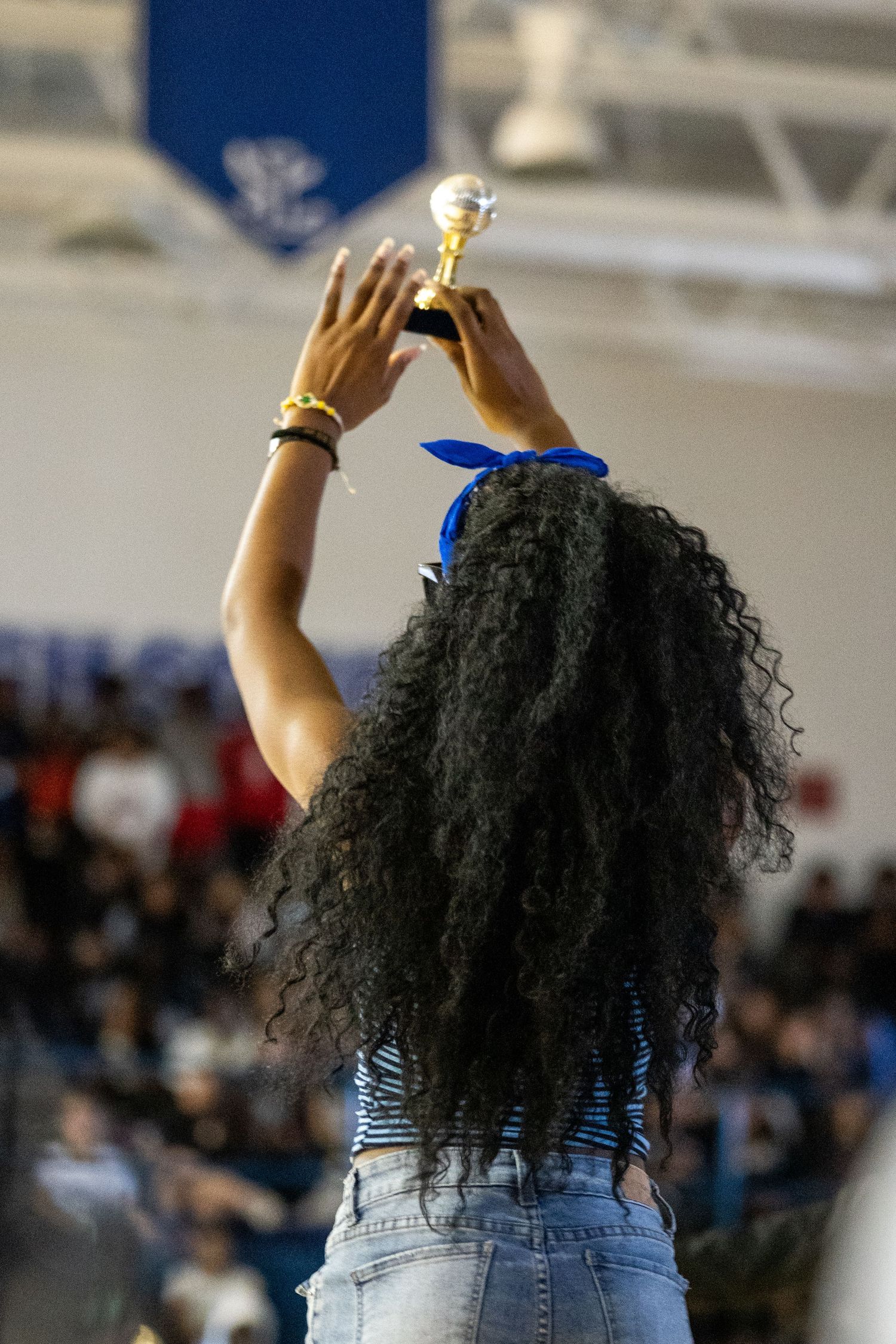 A young woman with curly hair raises a gold trophy above her head at a school gymnasium celebration event.