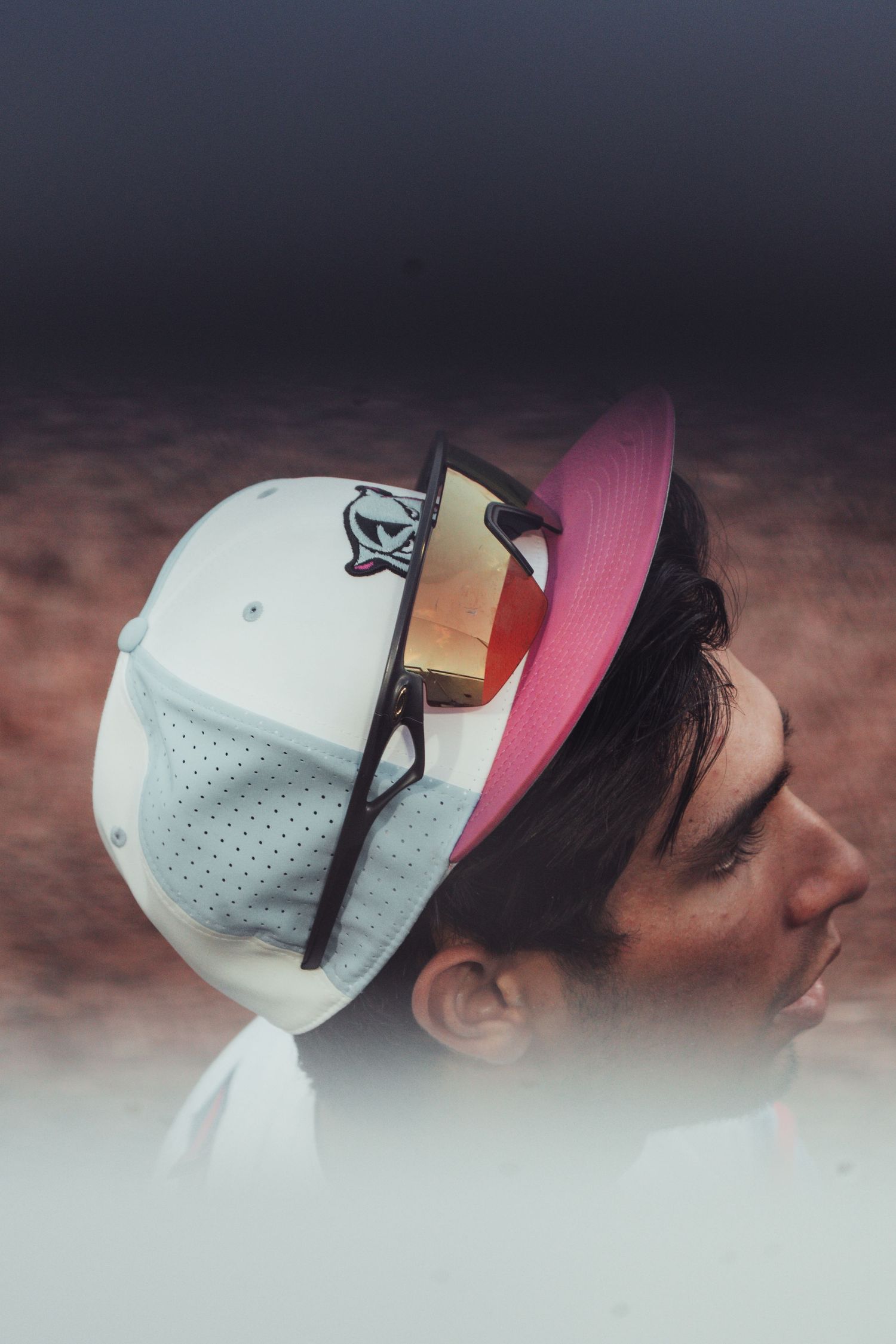 Young man in profile wearing stacked white and pink baseball caps with orange-tinted sunglasses perched on top.