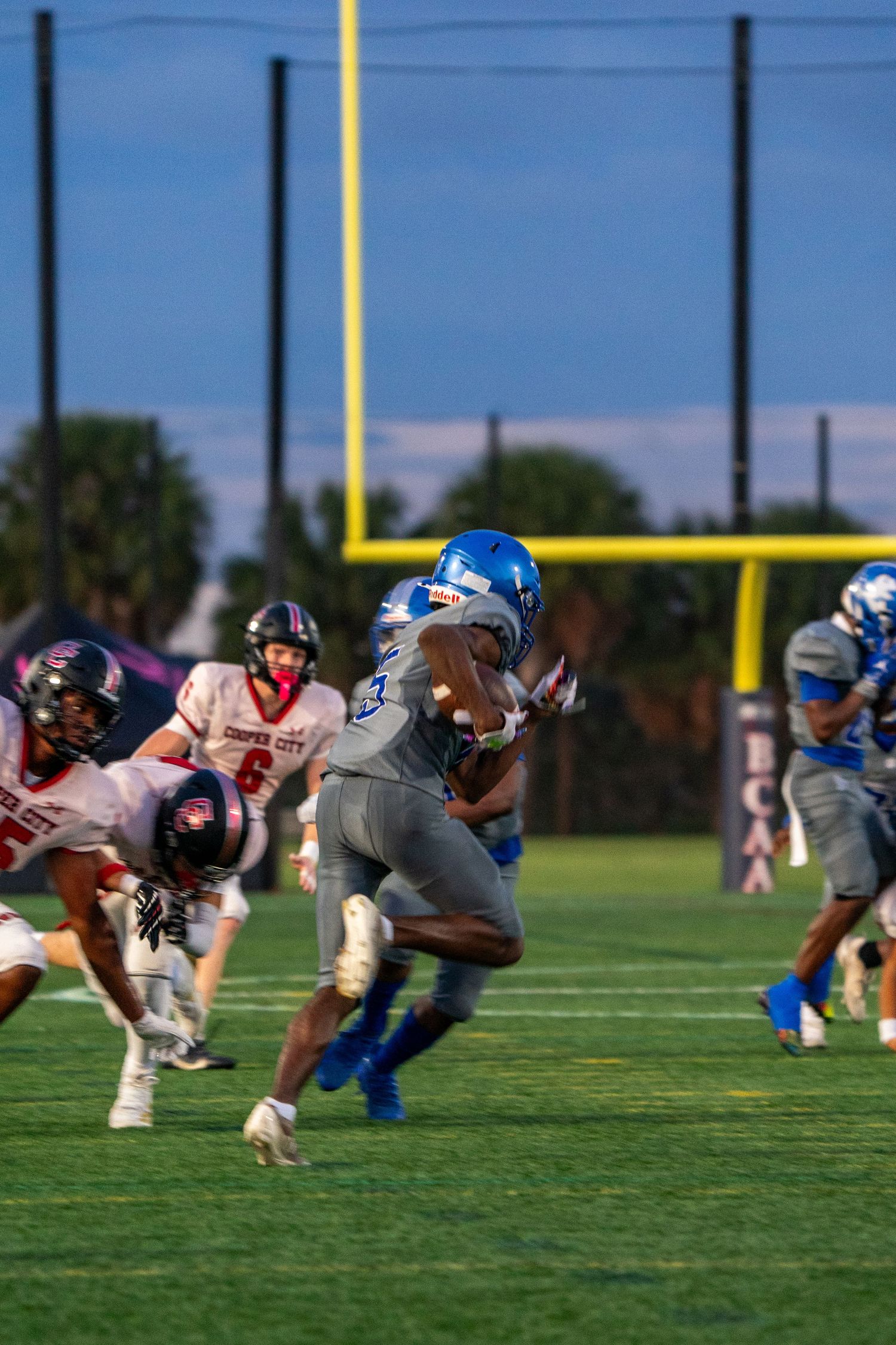 Football players line up in formation during practice on a sunny field with goal posts visible in the background.