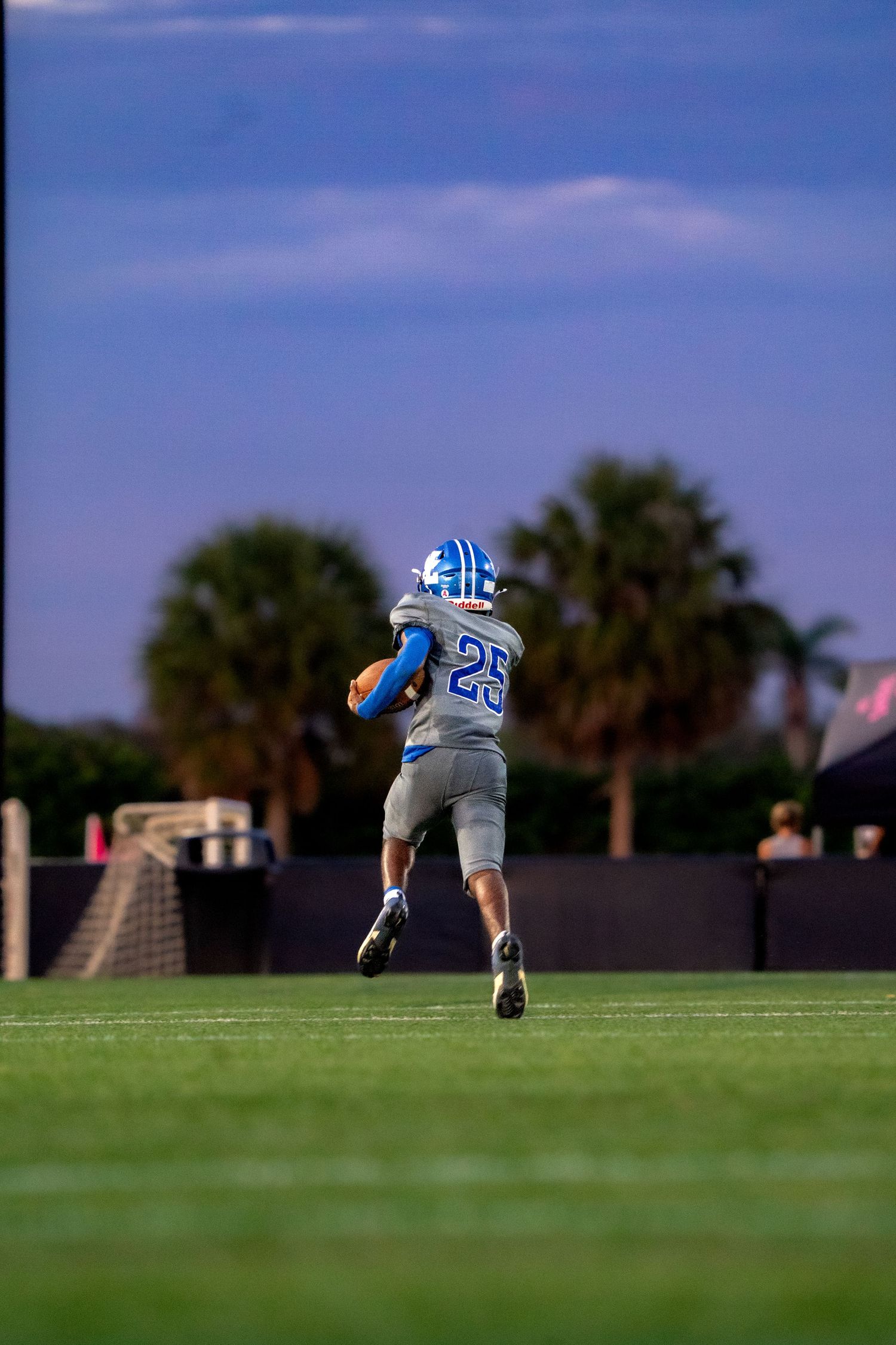 Player in light blue uniform diving on sports field with palm trees in background.