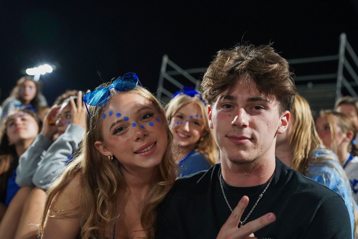 Students celebrating and making peace signs in crowded stadium bleachers at night.