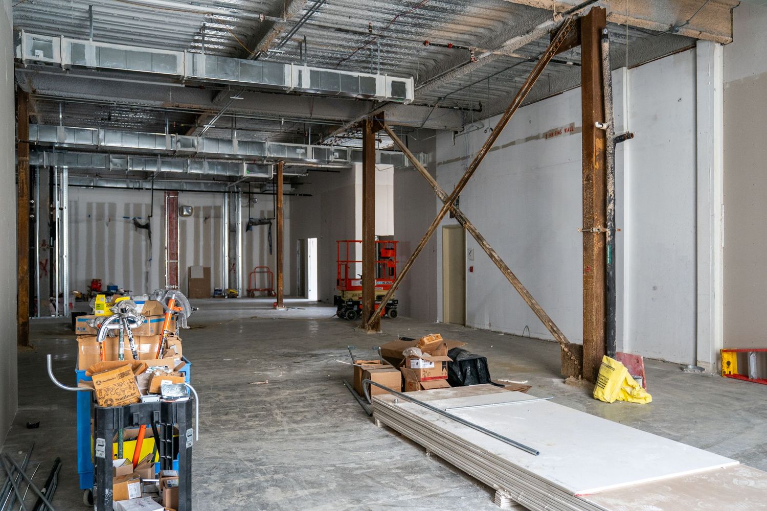 Construction site interior with exposed pipes, building materials, and orange lift equipment.