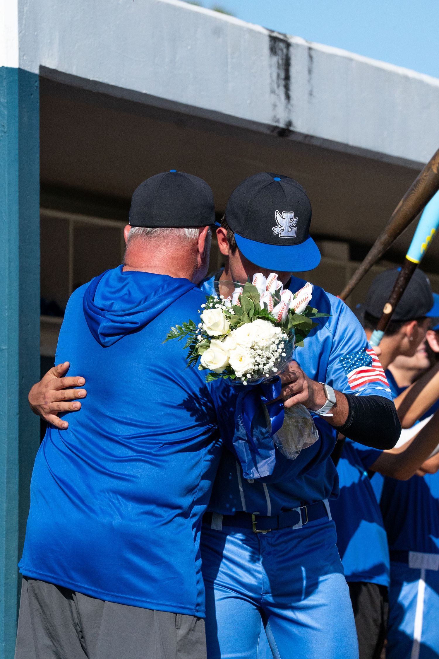Baseball player in blue uniform hugs coach while holding white flower bouquet during senior day ceremony.