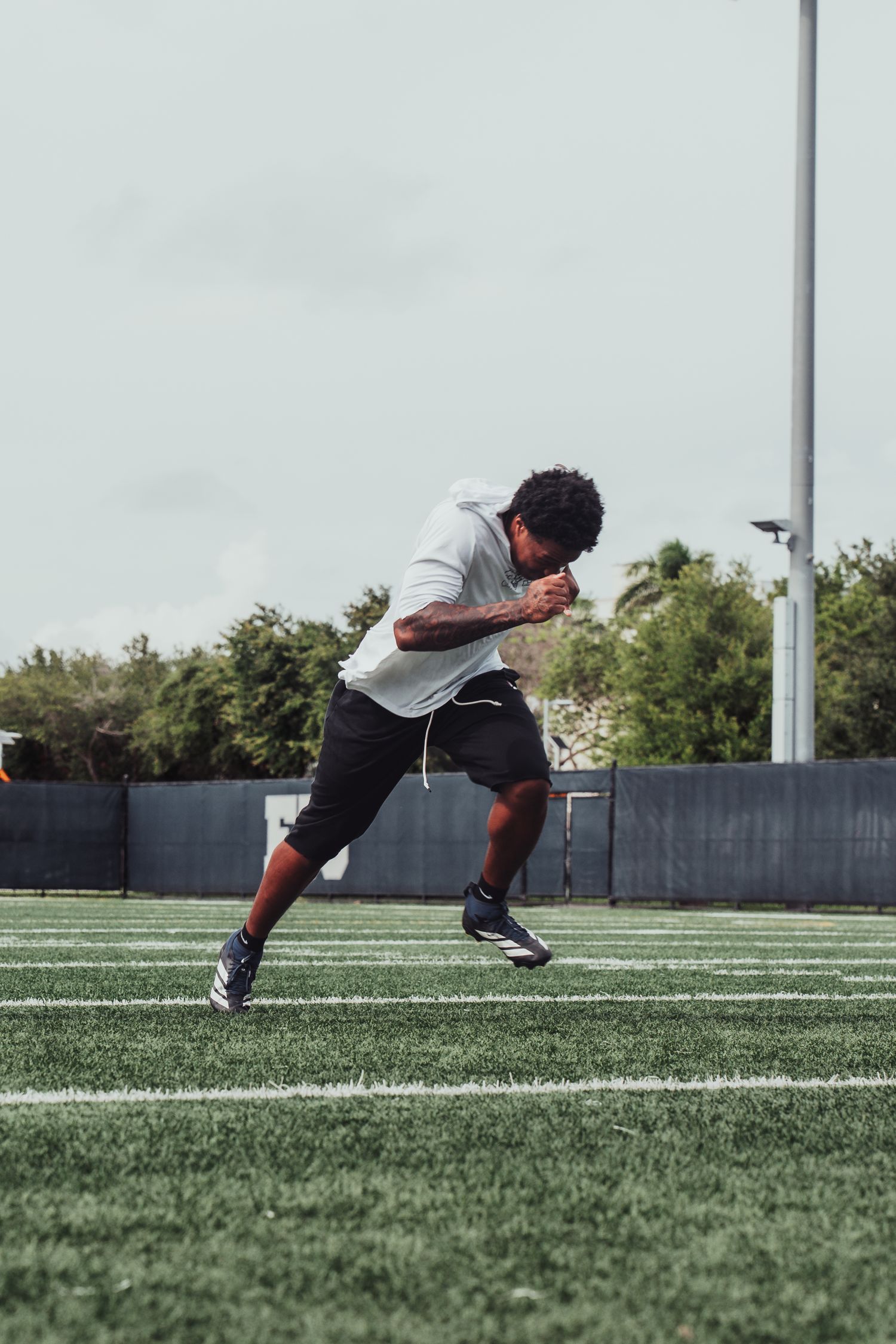 Athlete sprinting on green turf football field wearing white hoodie, black shorts, and cleats during outdoor training.