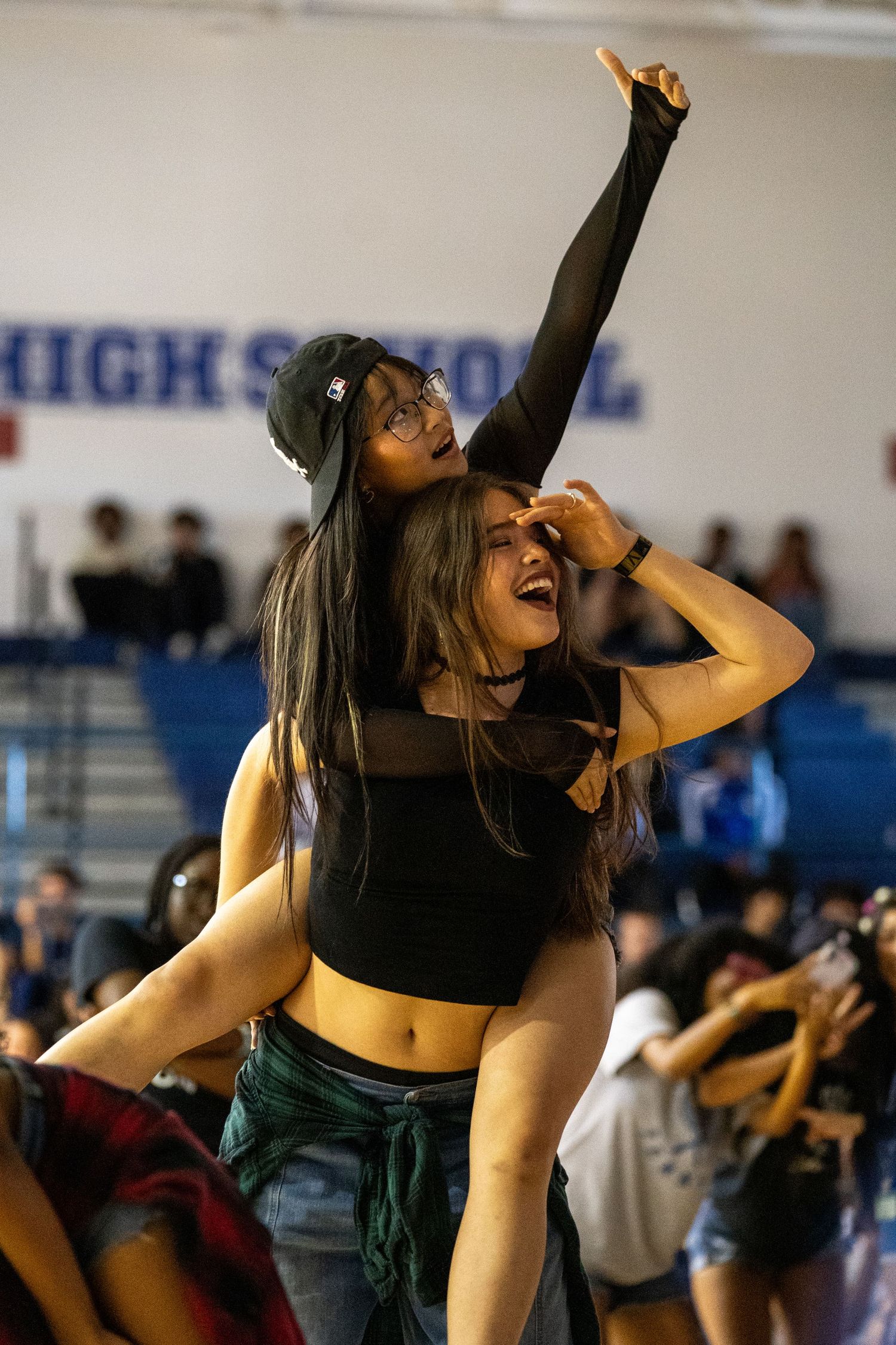 Two girls celebrate at a high school pep rally, one sitting on the other's shoulders with arm raised.
