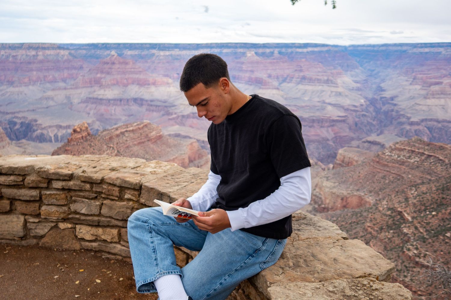 A person in casual attire reads while perched on a stone wall at the Grand Canyon.