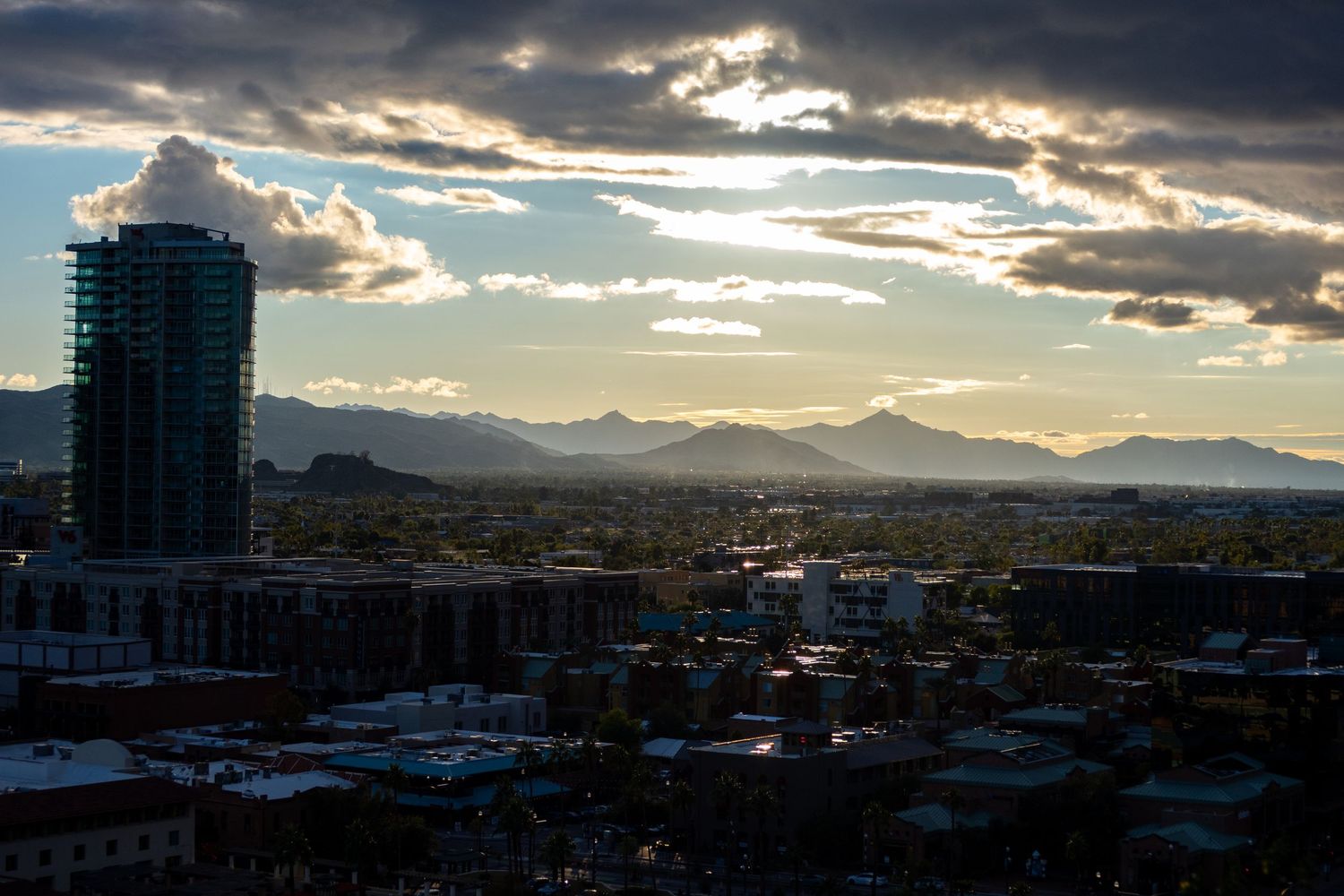 City buildings and mountains silhouetted against dramatic sunset sky with rays breaking through clouds.