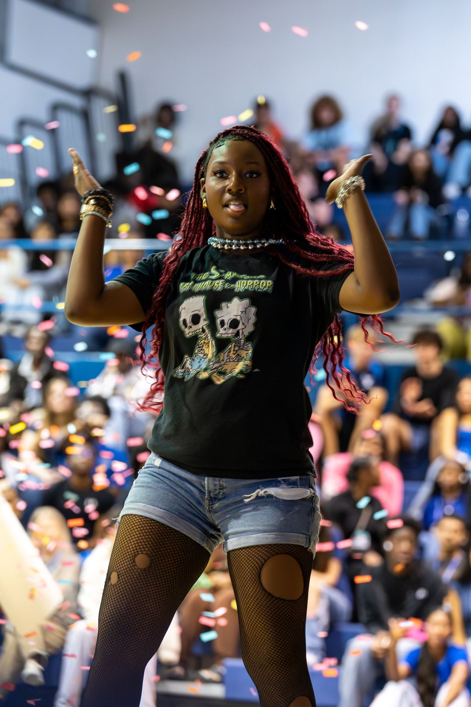 A young woman with red braids dances in fishnets and skull tee amid colorful confetti at a lively indoor event.