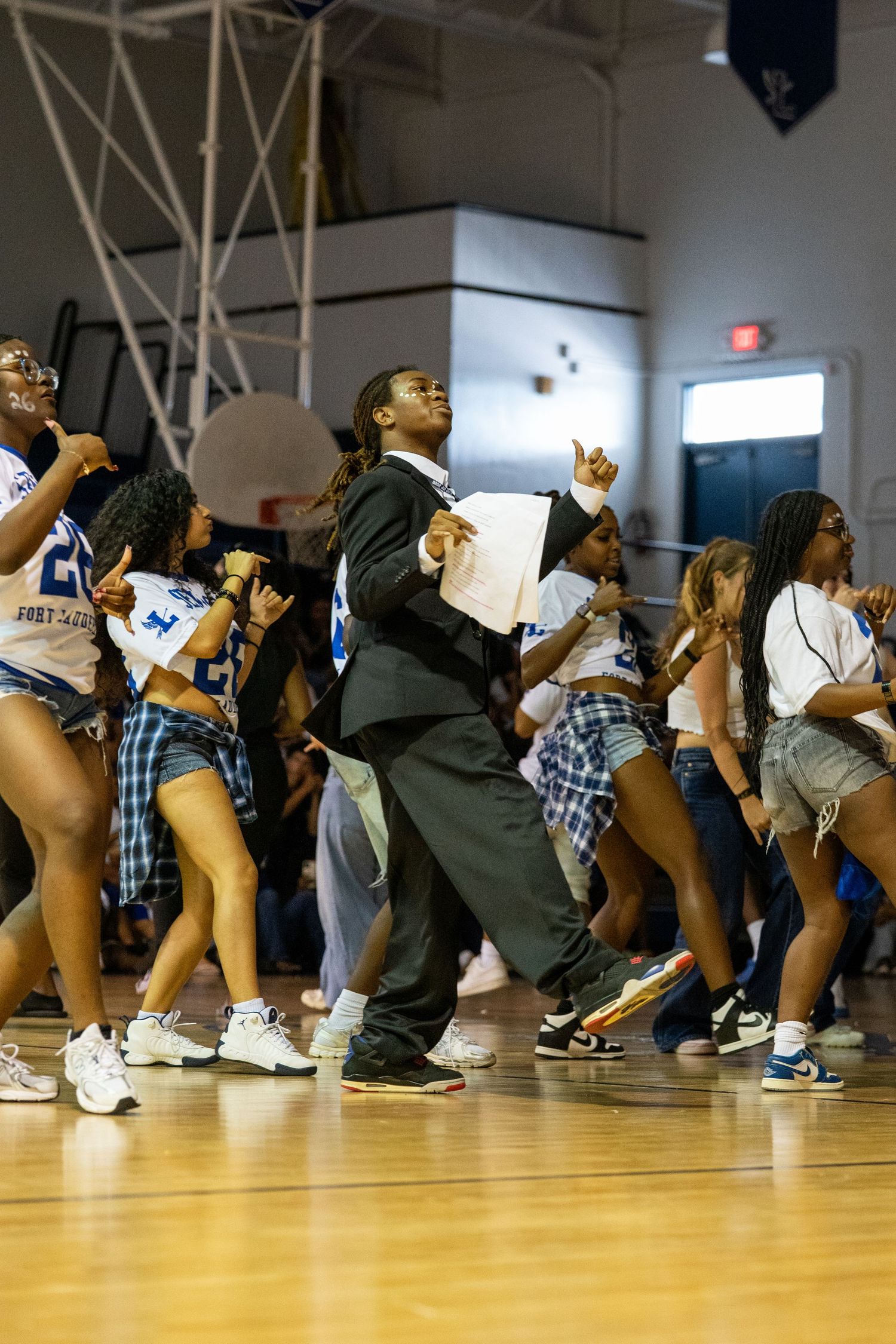 Students dancing energetically in a school gymnasium during a pep rally, wearing blue and white outfits.