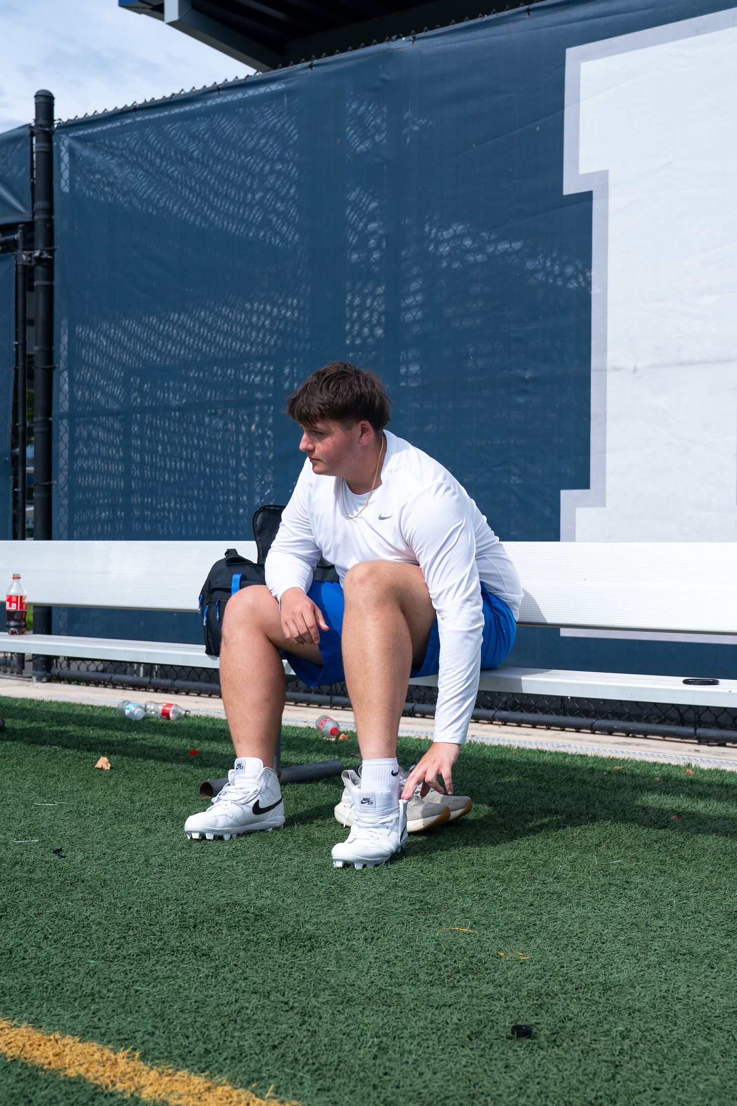 Young athlete in white Nike gear sits on a bench on turf field, resting during a sports practice session.