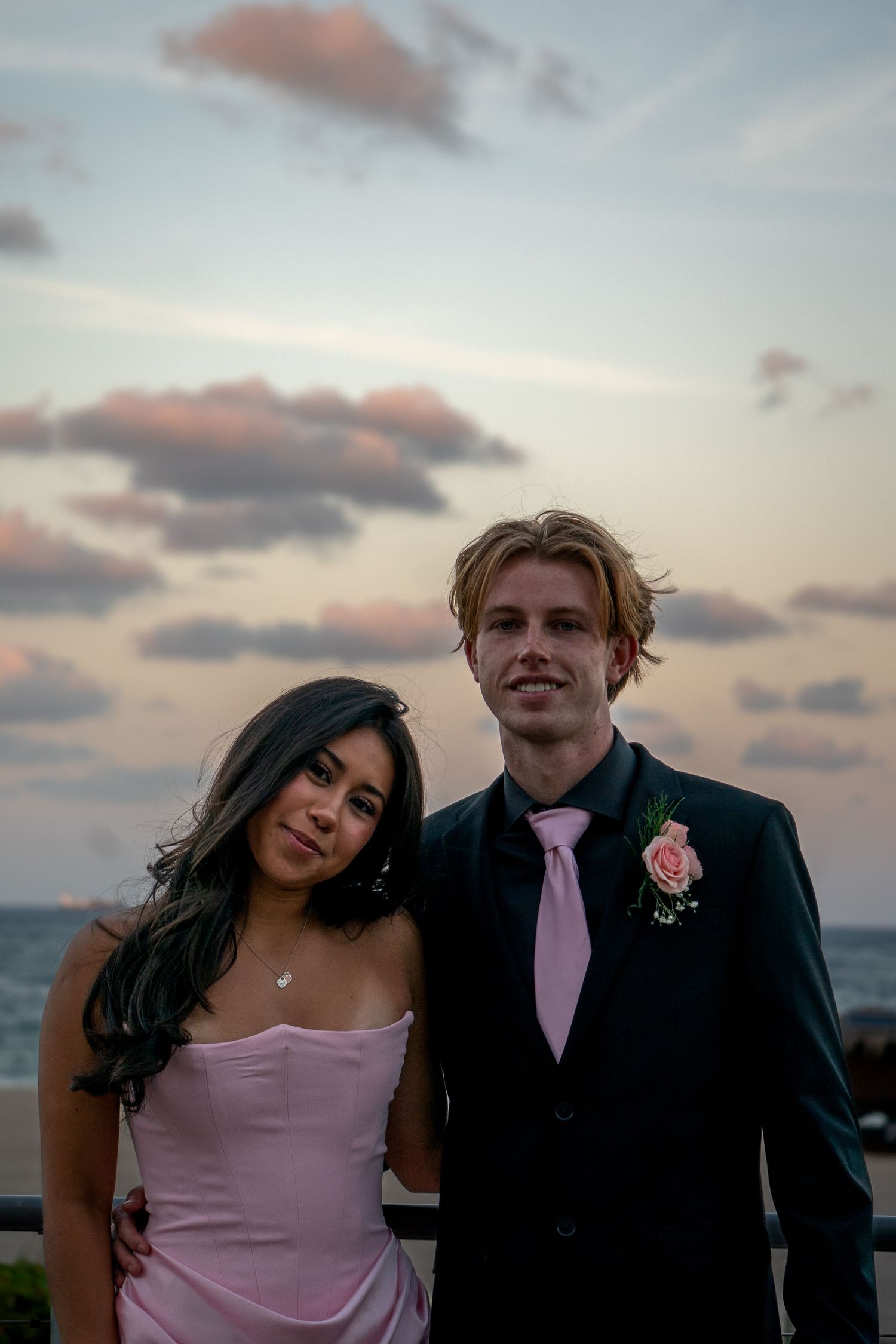 Couple in formal attire posing against sunset sky at outdoor event.
