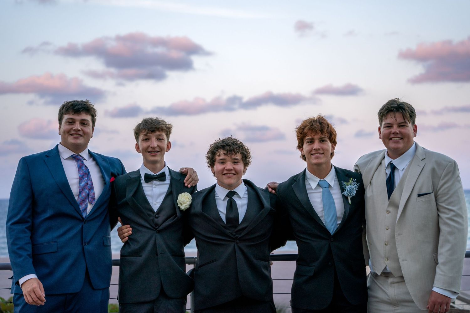 Group wearing formal attire poses together against sunset sky at outdoor event.