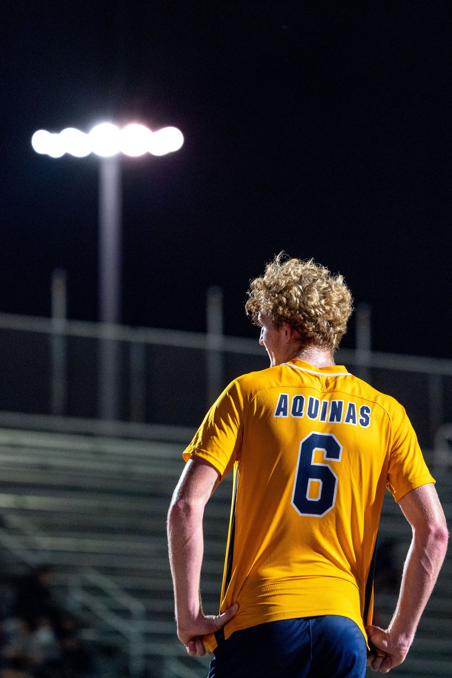 Soccer player in yellow Aquinas jersey number 6 standing under stadium lights.