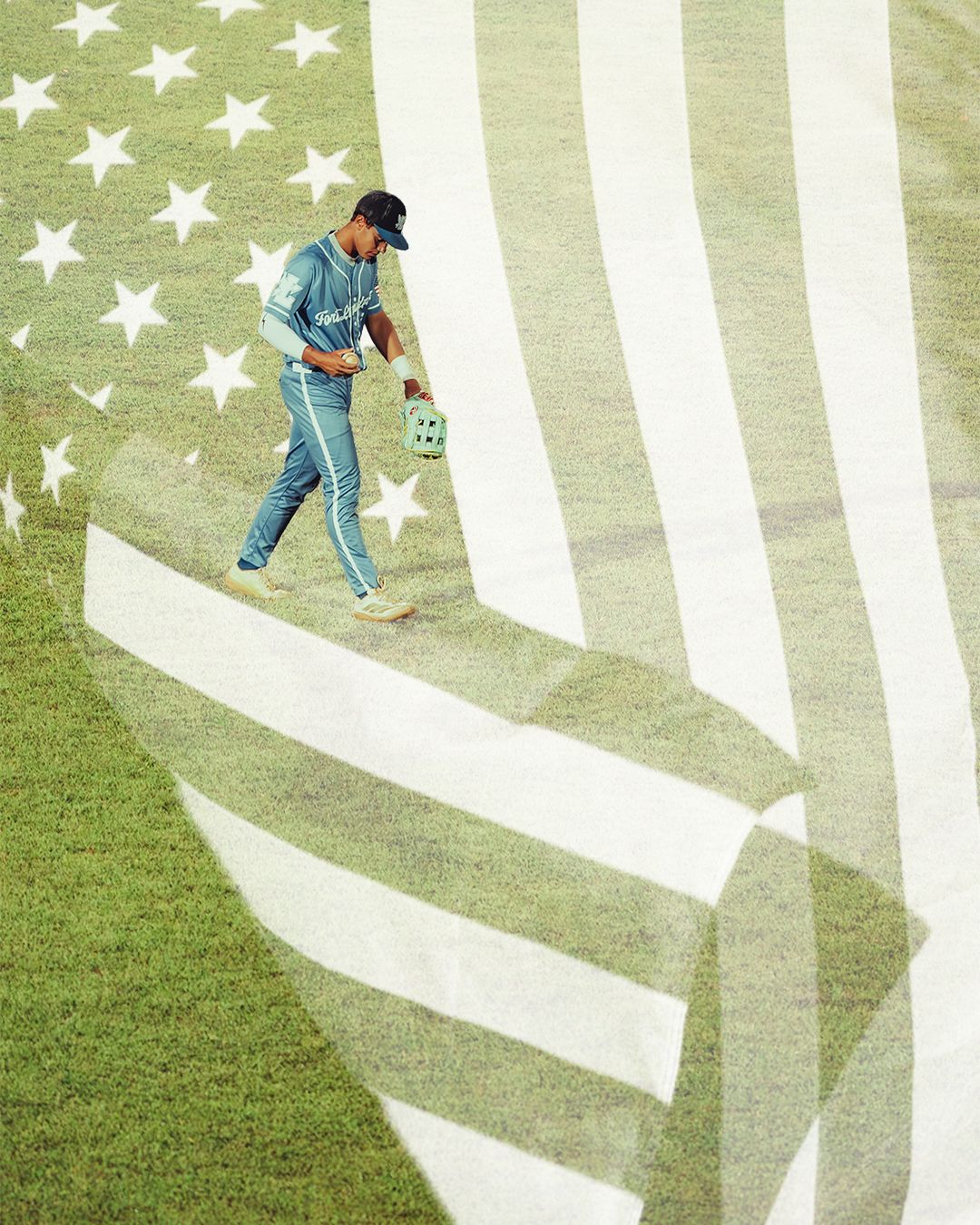 Baseball groundskeeper walks across a large American flag painted on green stadium grass.