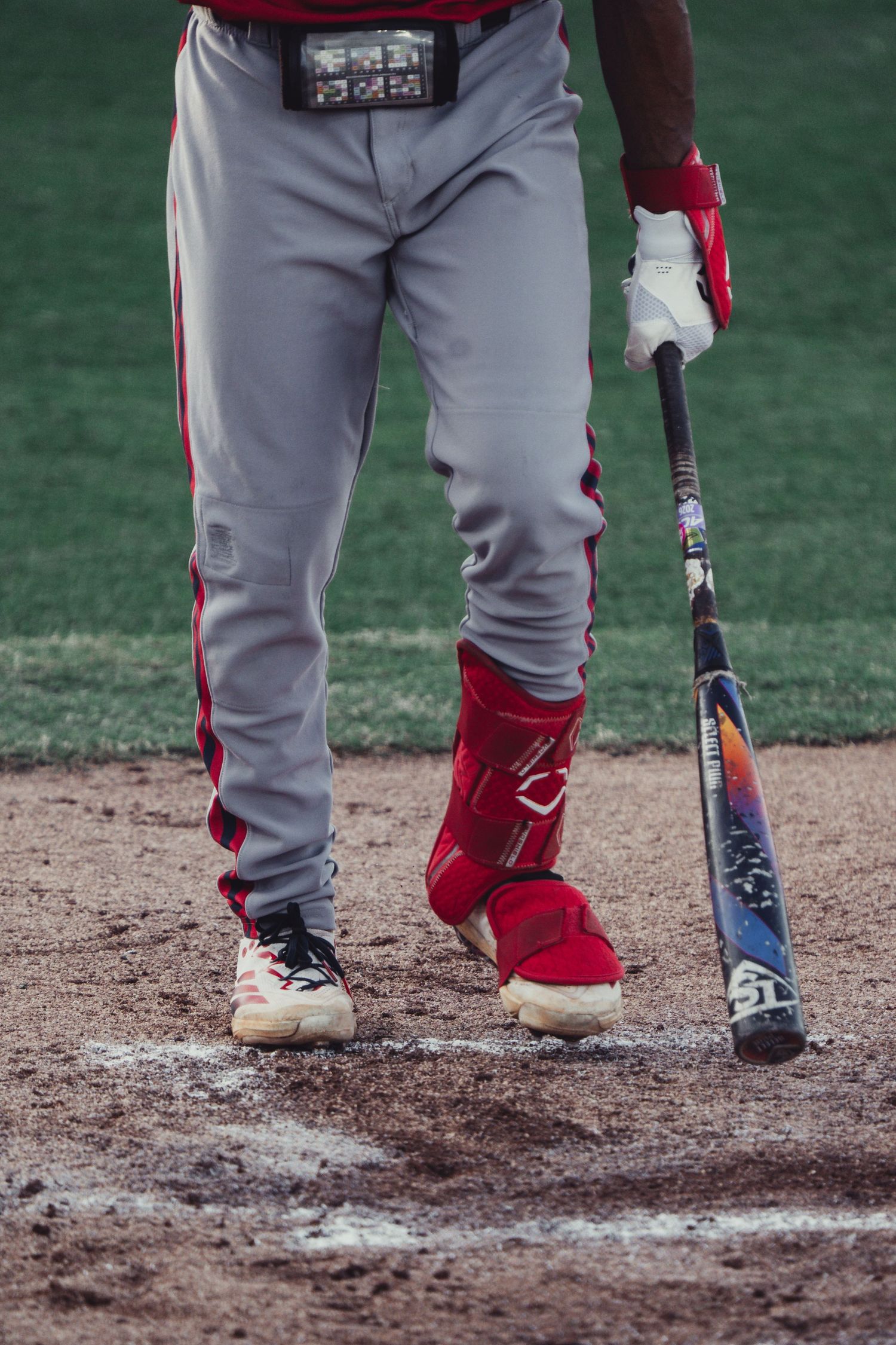 Baseball player in gray and red uniform holding a bat at home plate, wearing red ankle guard and cleats.