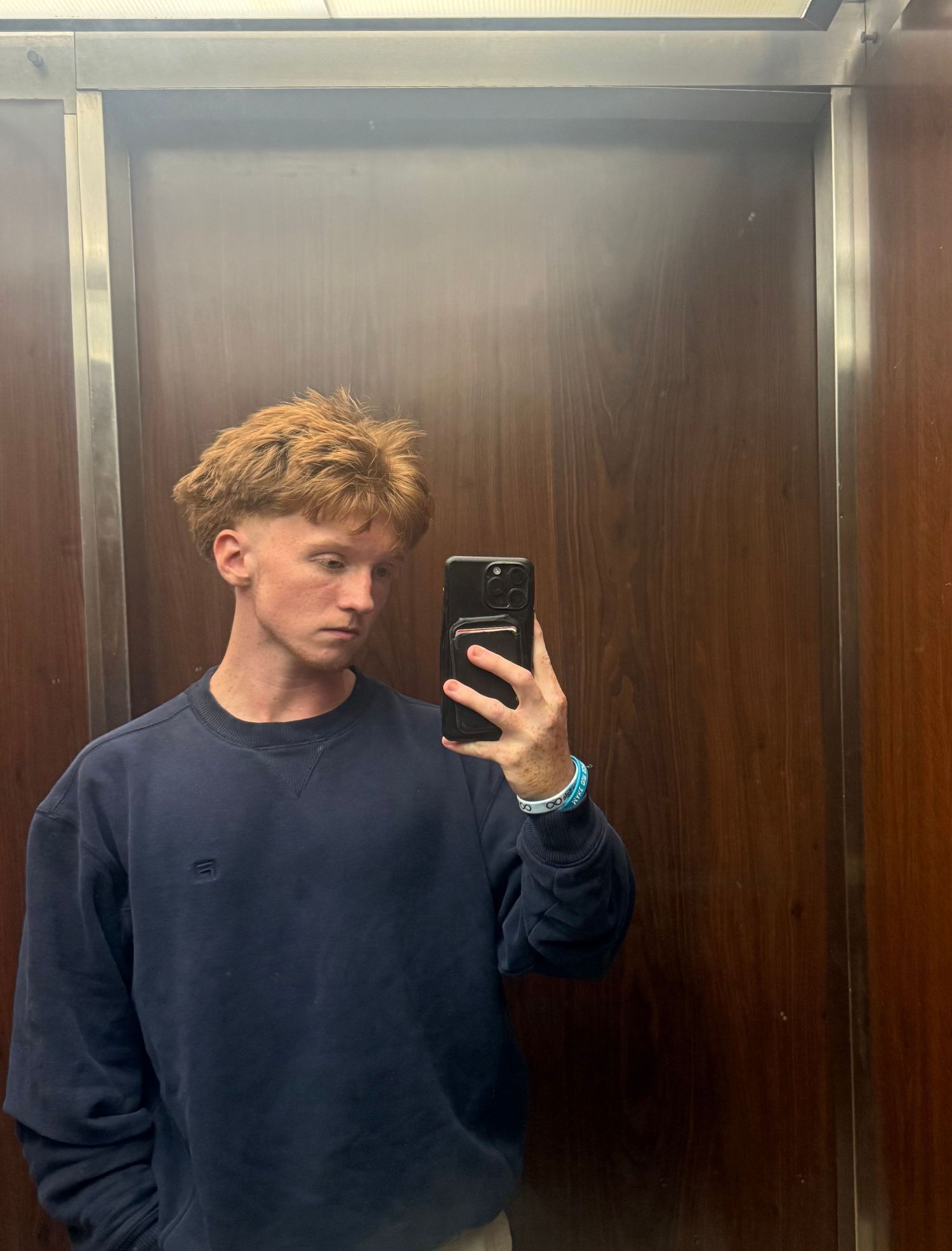 A person wearing a navy blue sweatshirt takes mirror selfies in an elevator with wood paneled walls.