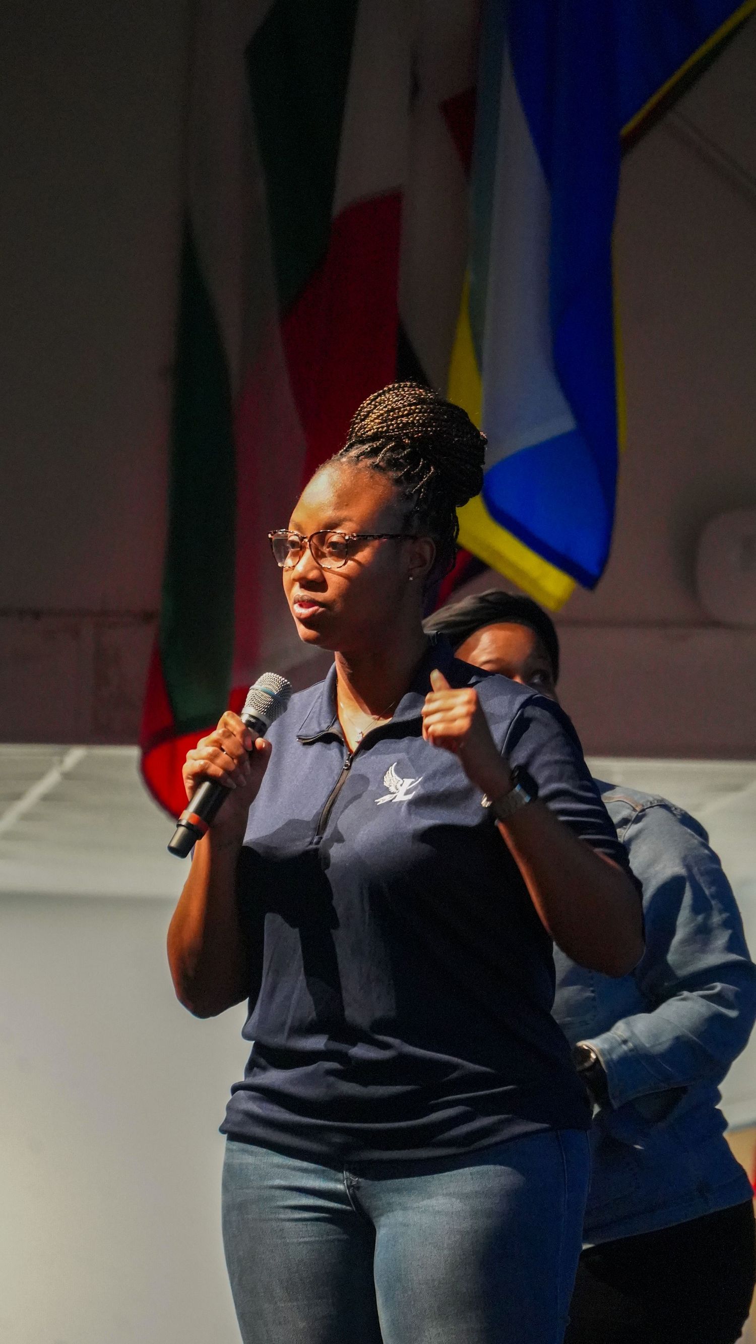 A performer in navy uniform speaks into microphone with flags in background.