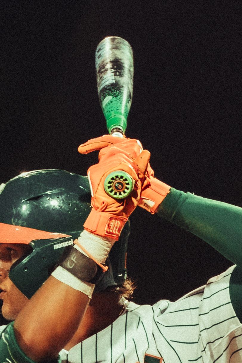 Baseball batter in orange gloves grips a green bat, wearing a black helmet under bright stadium lights at night.