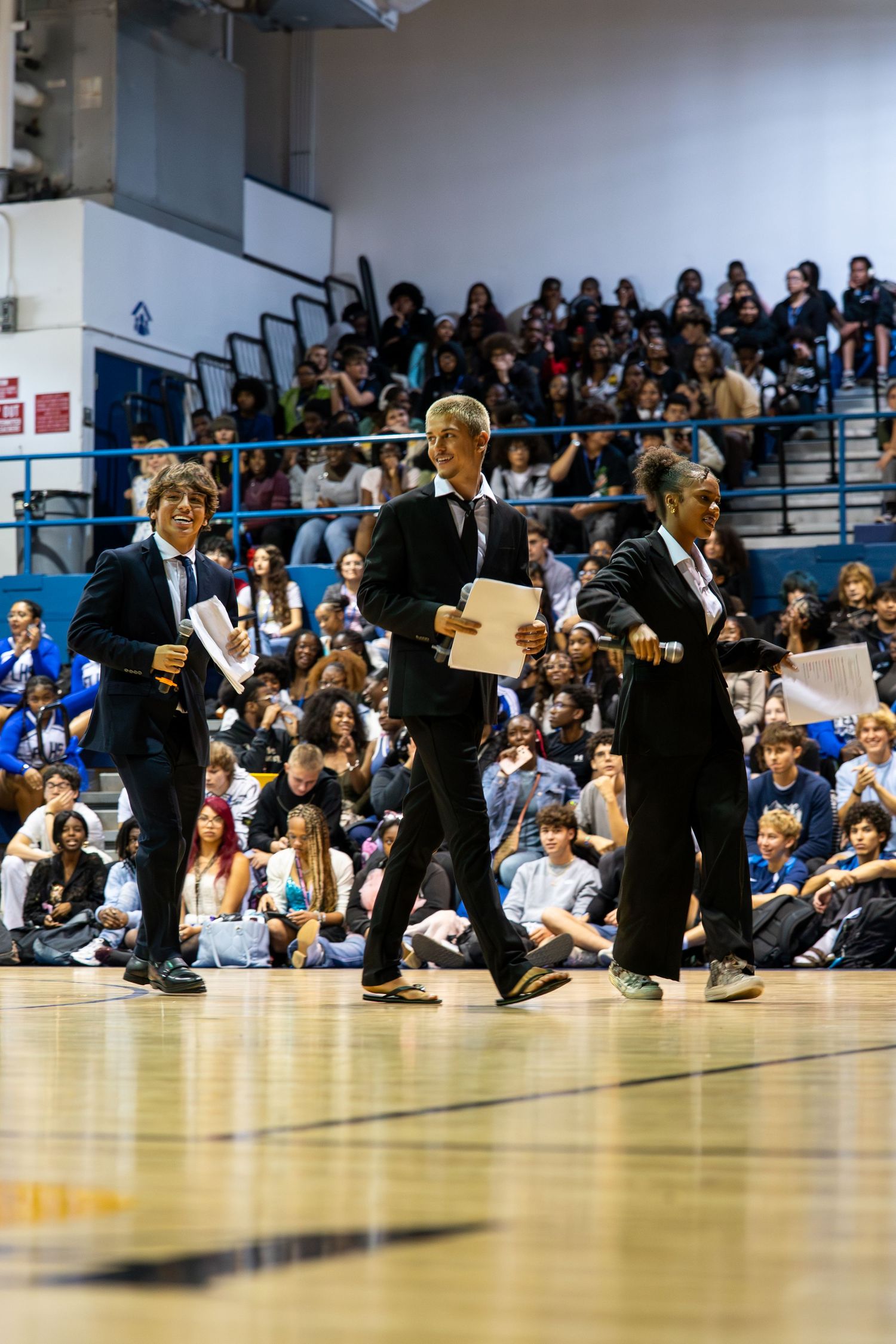 Three students in formal attire walk across a school gymnasium floor during a pep rally assembly.