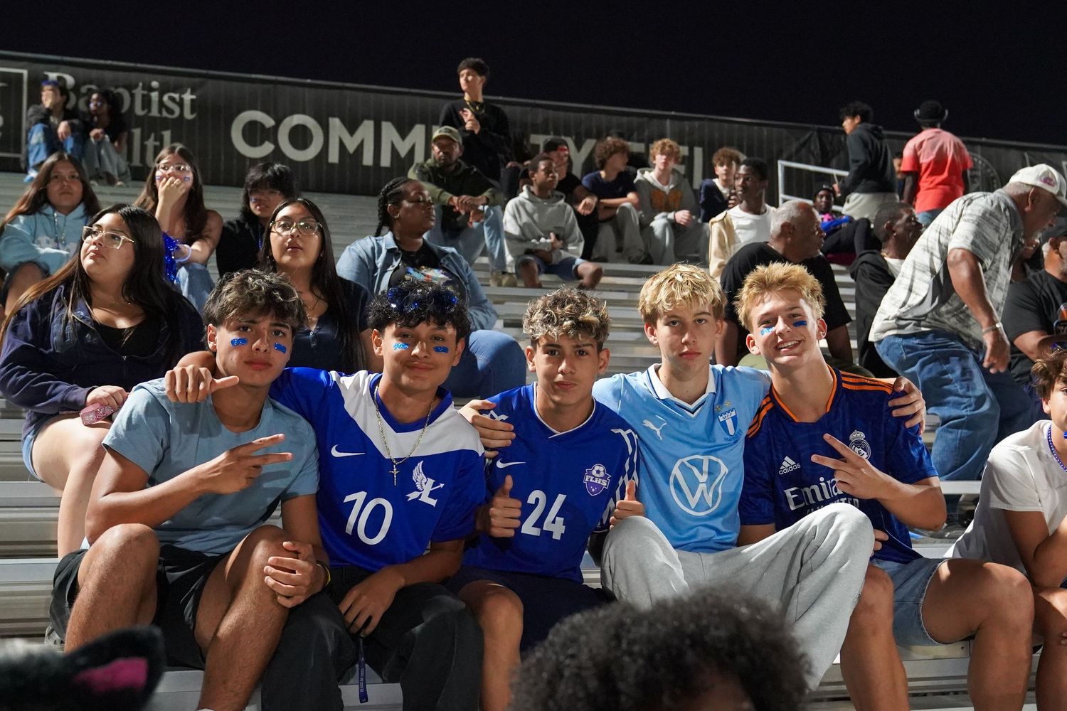 Students in blue jerseys watching intently from stadium bleachers during a nighttime game.