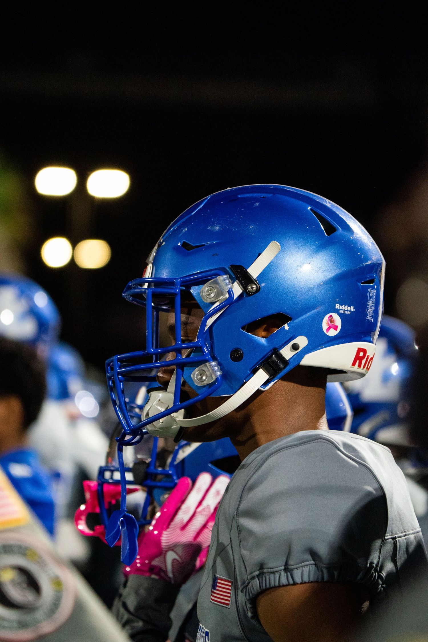 Row of blue football helmets with pink accents lined up on the sideline.